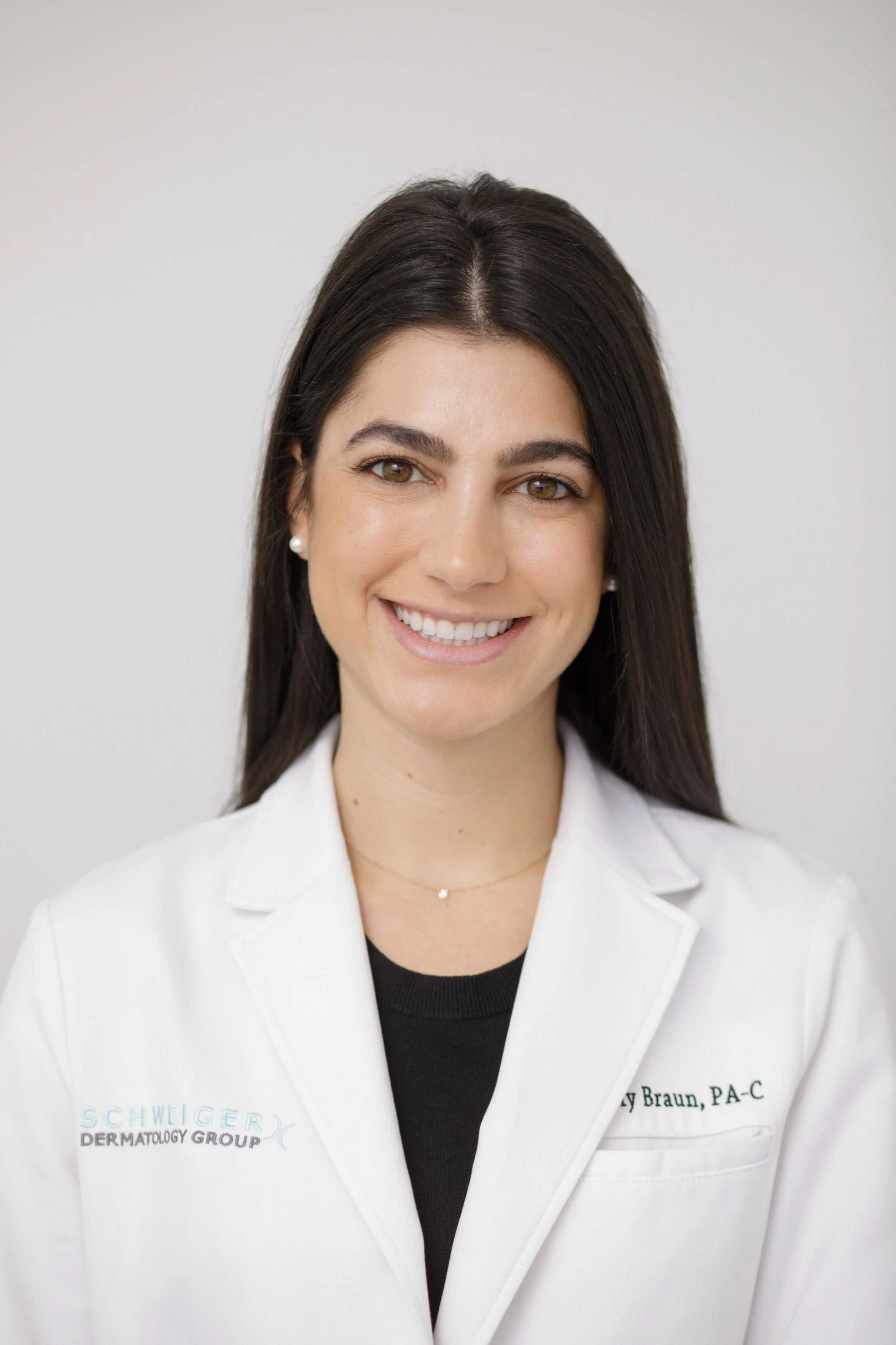 Professional woman with long dark hair, wearing a white coat with the text 'Schweiger Dermatology Group' and 'Ly Braum, PA-C', smiling against a plain background.
