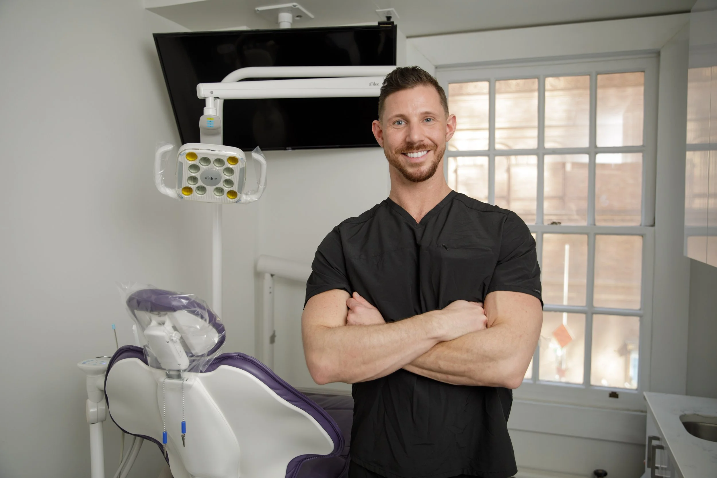 A smiling male dentist standing in a dental clinic with arms crossed, next to a dental chair and equipment, with a window in the background.