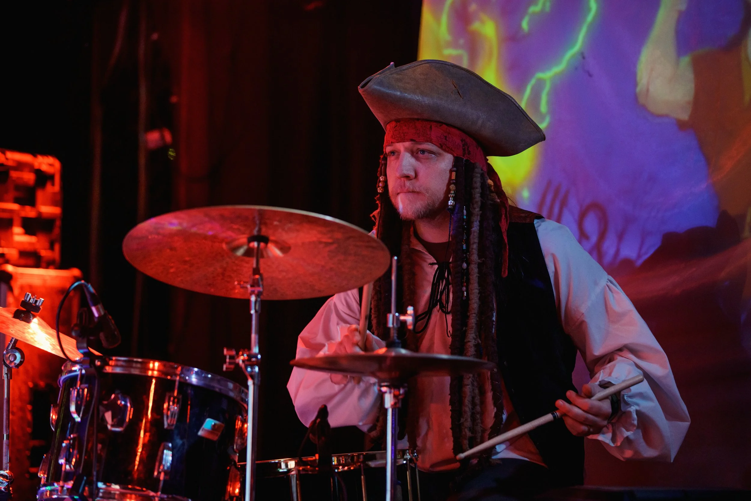 A man dressed as a pirate with a large pirate hat, red bandana, and dreadlocks, playing drums on stage with colorful lighting and a vibrant backdrop.