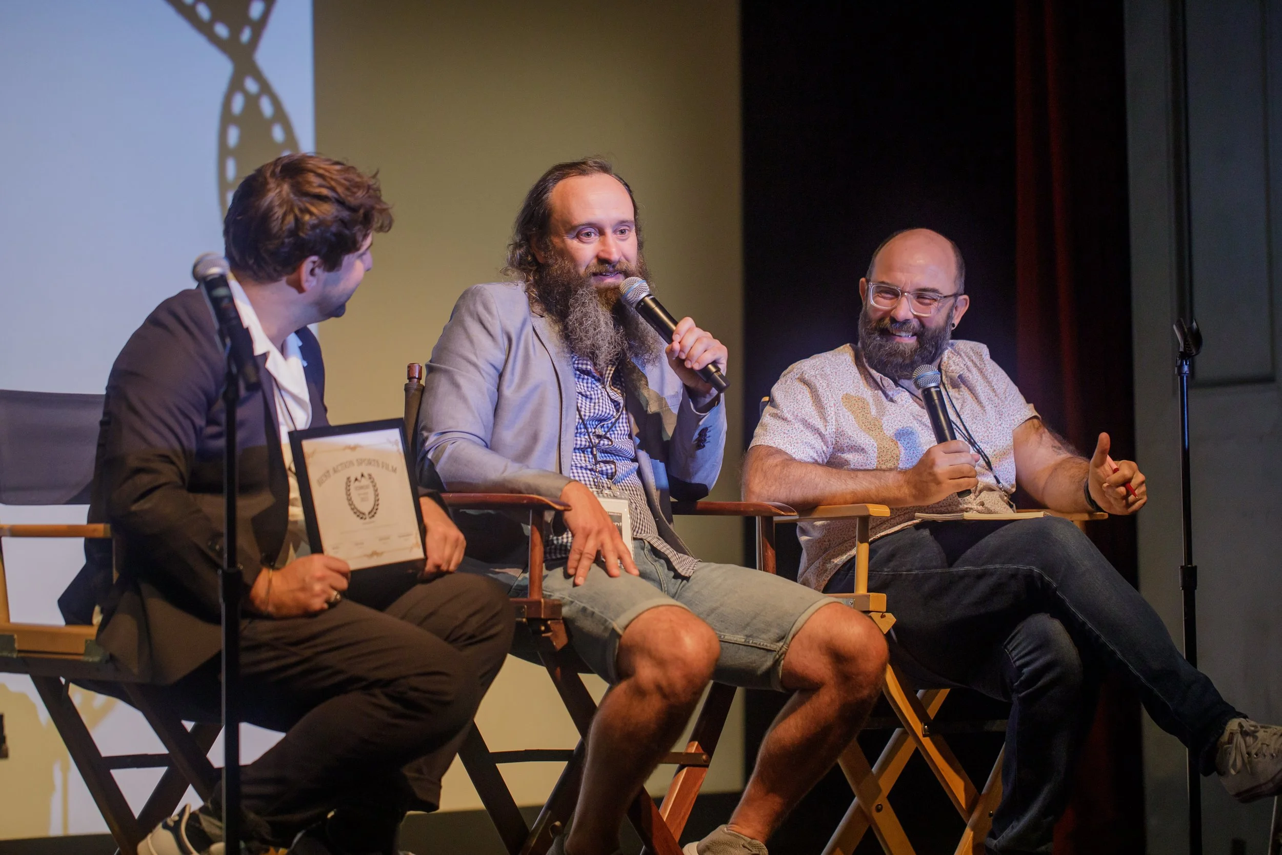 Three men seated on stage at a panel discussion, holding microphones and a framed award, with a projection screen behind them.