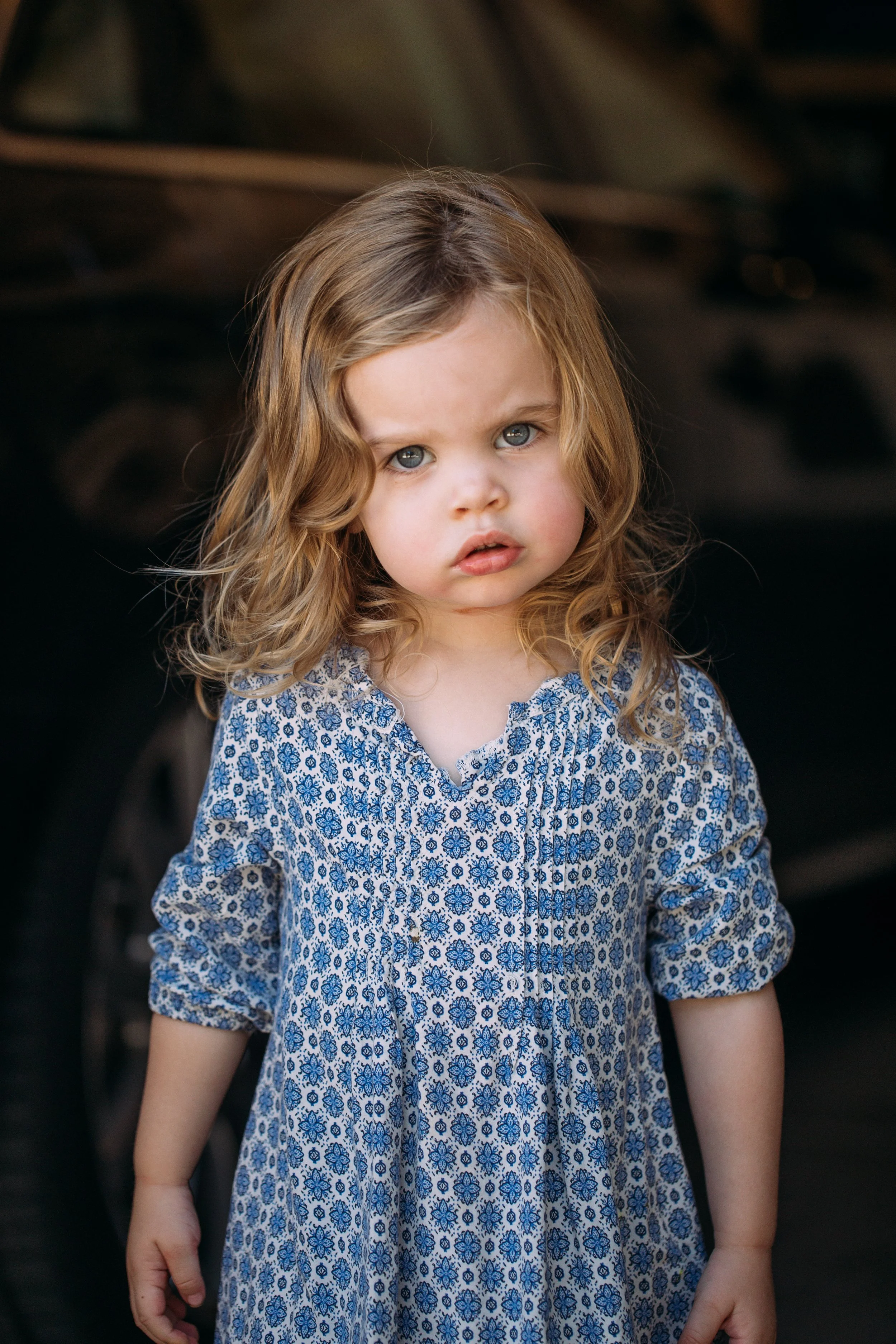 A young girl with wavy brown hair and blue eyes, wearing a blue and white patterned dress, stands outdoors against a dark background.