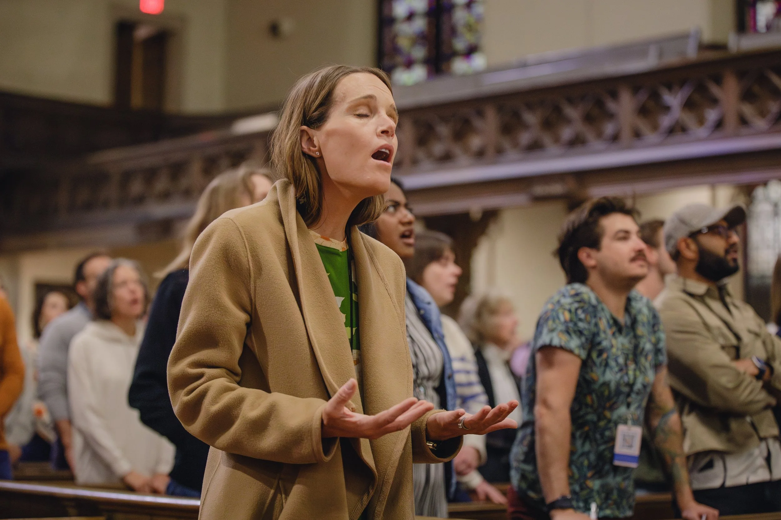 A woman with shoulder-length hair and closed eyes, standing with her hands clasped, appears to be singing or praying in a church or concert hall, surrounded by a diverse group of people who are also engaged in the same activity.