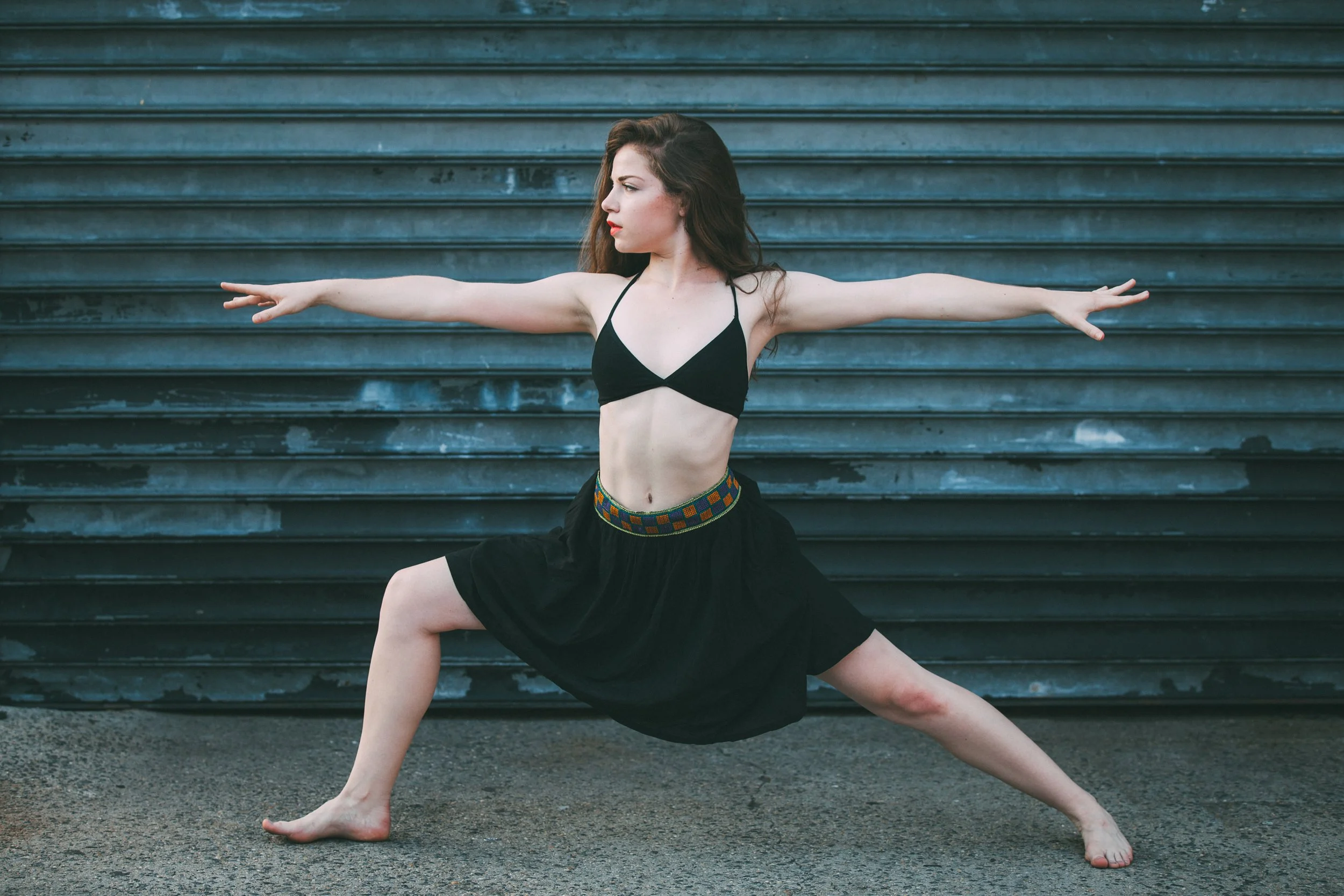 A woman with long brown hair in a yoga pose outdoors in front of a metal staircase, wearing a black sports bra and black skirt with a colorful waistband.