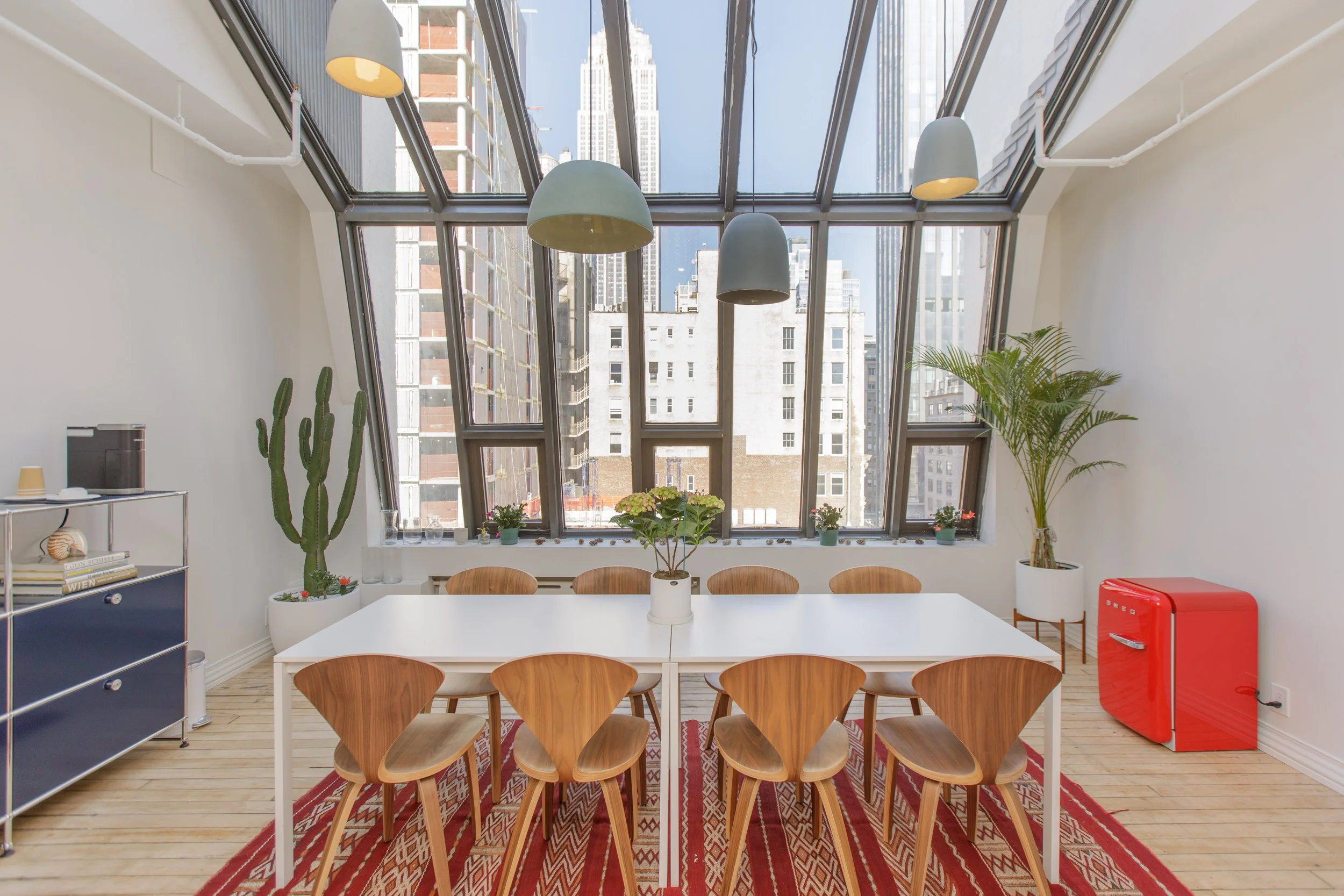 A modern dining room with a white table surrounded by wooden chairs, large angled windows with city views, and several potted plants. There are three hanging lights above the table, a patterned rug, a blue cabinet with books and decor, a red mini fri
