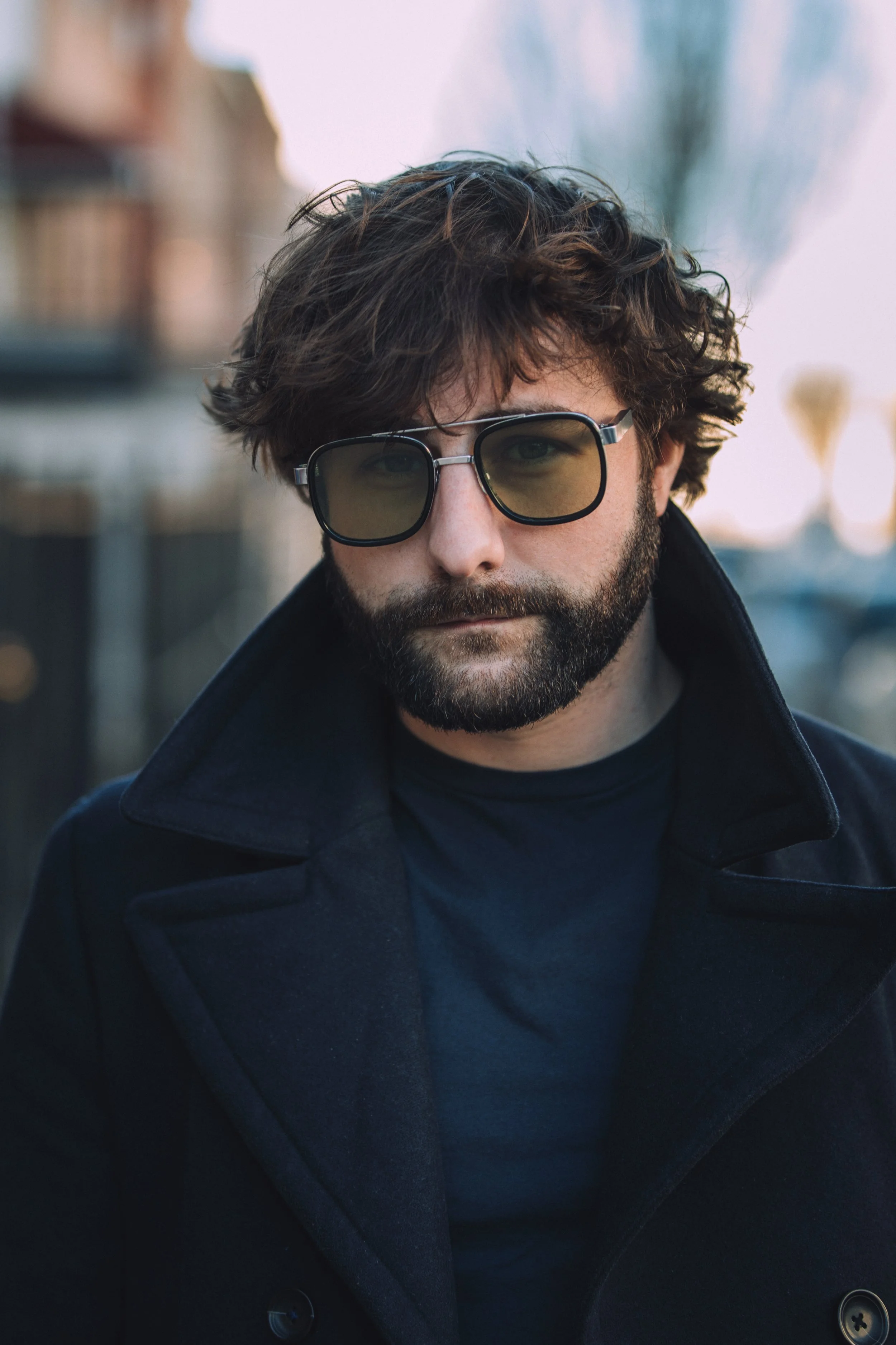 Close-up of a man with aquamarine eyes and a beard, wearing dark tinted glasses and a black coat outdoors with blurred buildings and trees in the background.
