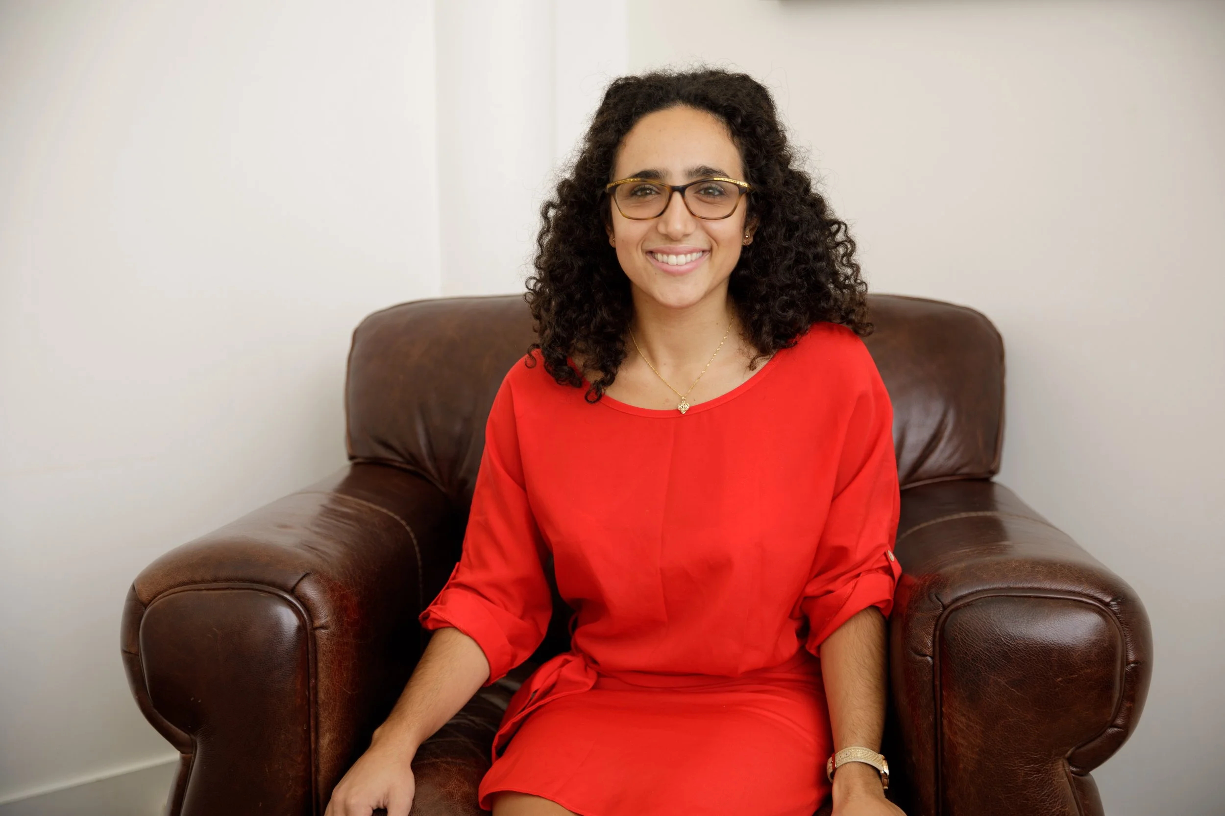 A woman with curly dark hair wearing glasses and a red dress, sitting on a brown leather armchair in front of a plain white wall, smiling at the camera.