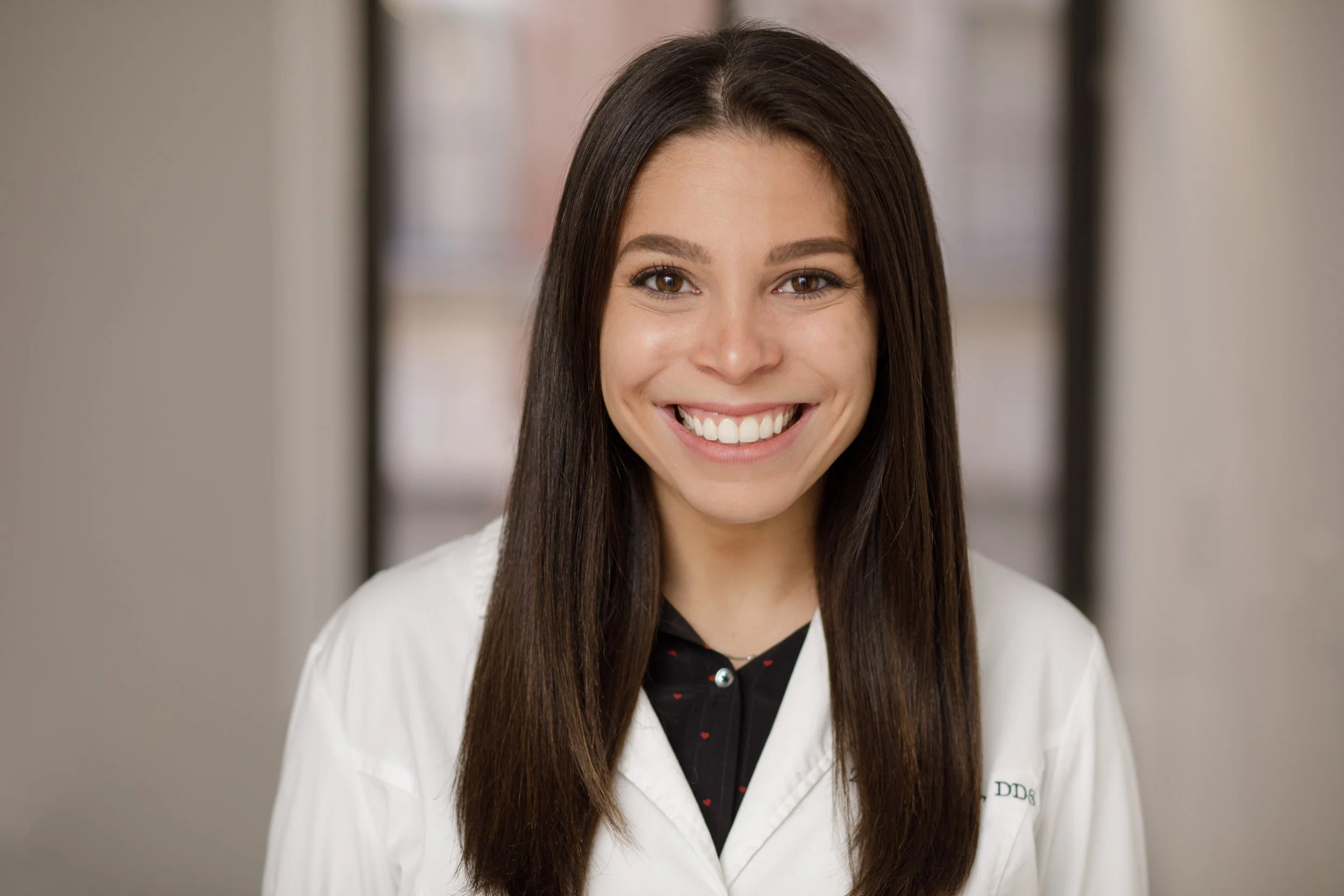 A smiling woman with long brown hair in a white lab coat, standing in a hallway with blurred background.