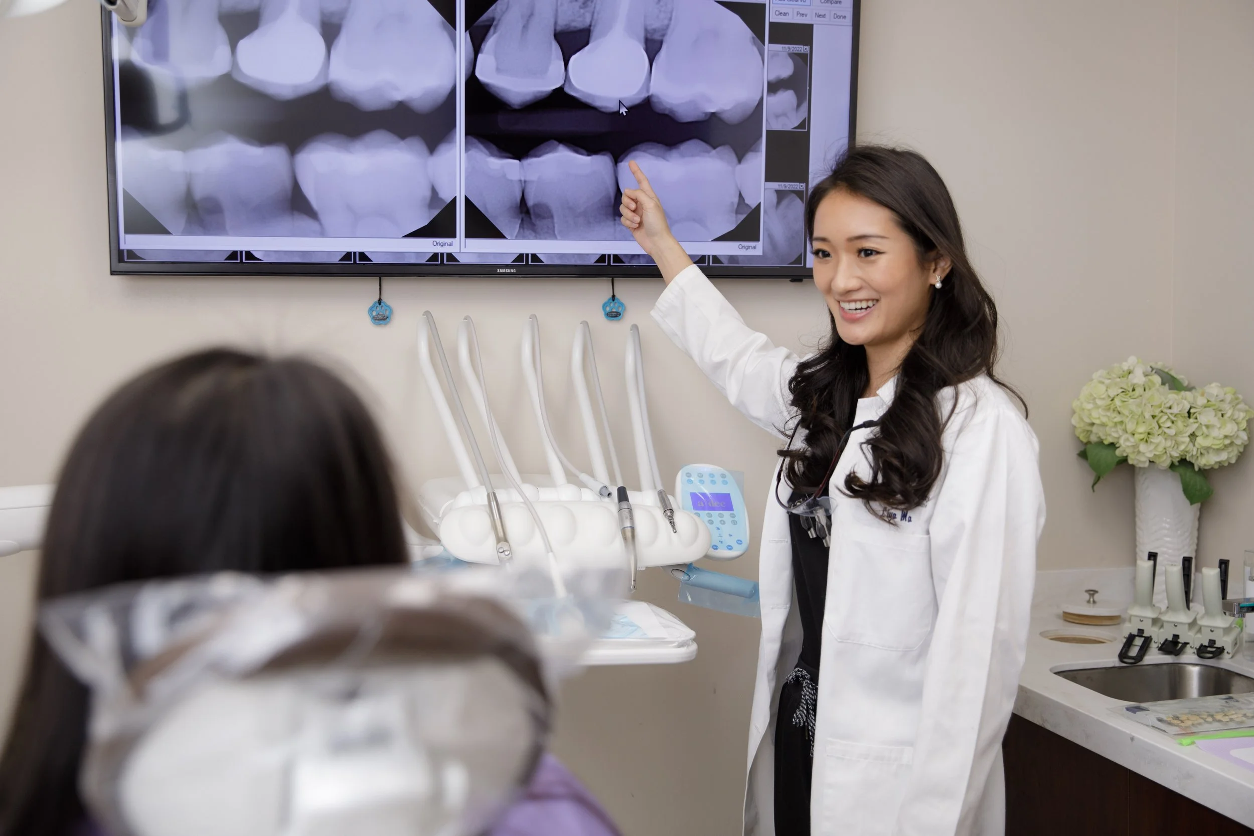 A female dentist smiling and pointing at dental X-rays on a monitor while talking to a patient in a dental office.