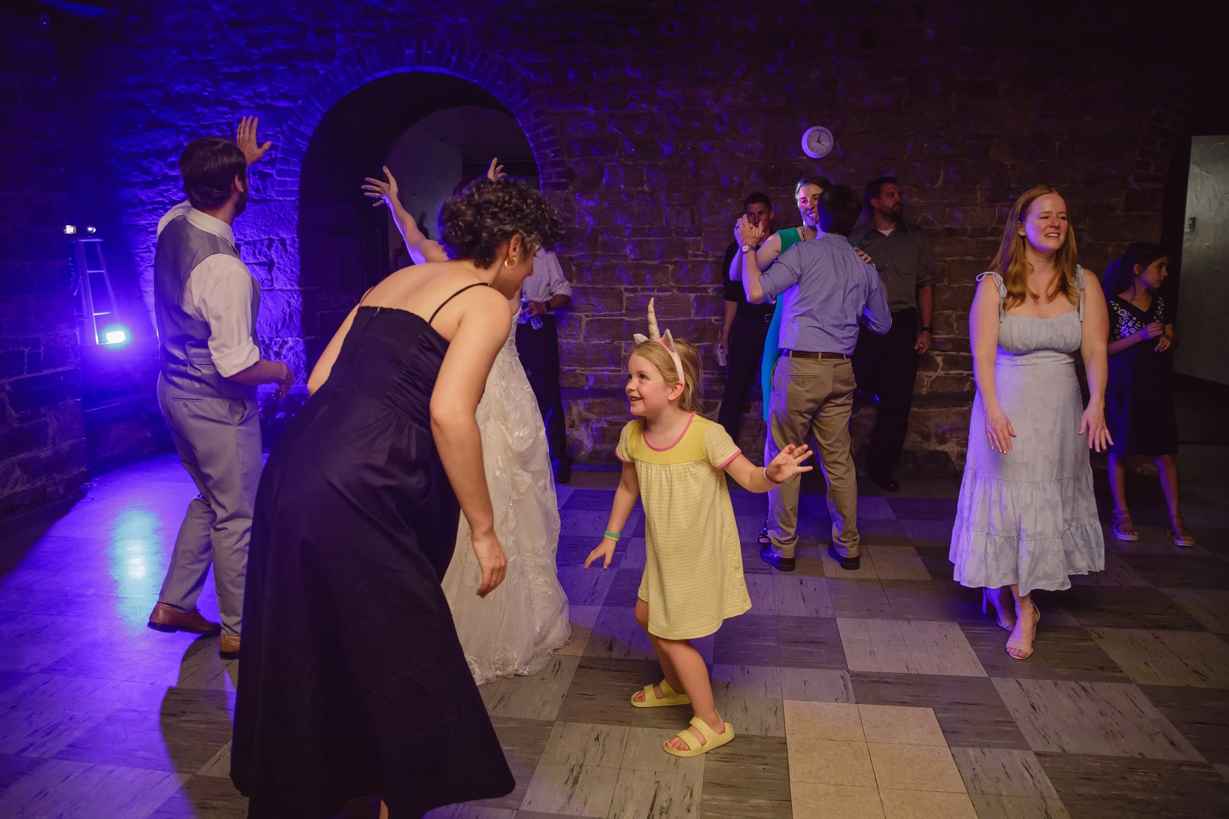 People dancing and socializing at a party with purple lighting and stone walls.