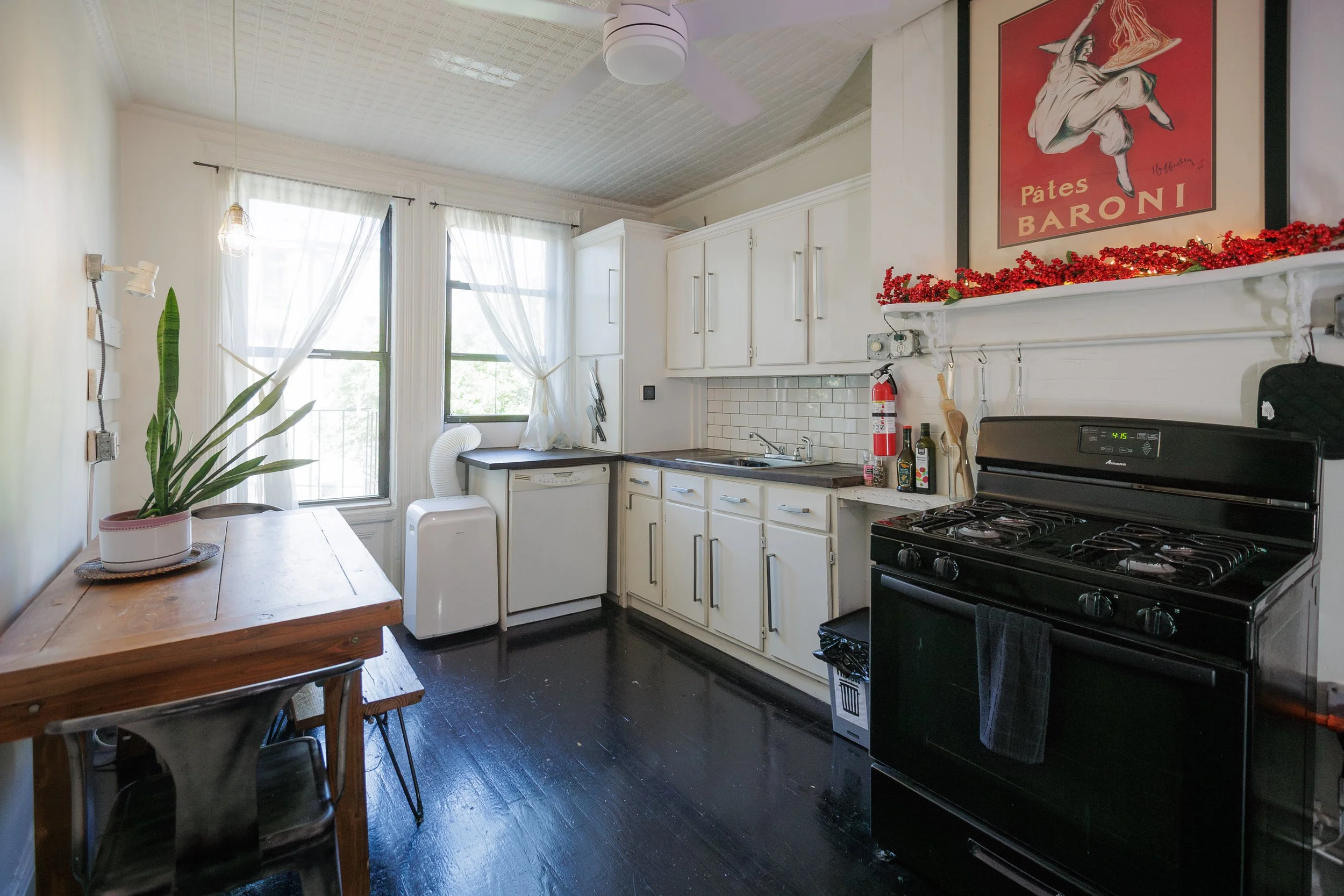 A kitchen with white cabinets, a black stove, a dishwasher, and a stainless steel sink. There are windows with white curtains, a wooden dining table with black chairs, a potted plant, and a decorative red berry garland above the white mantel with a l