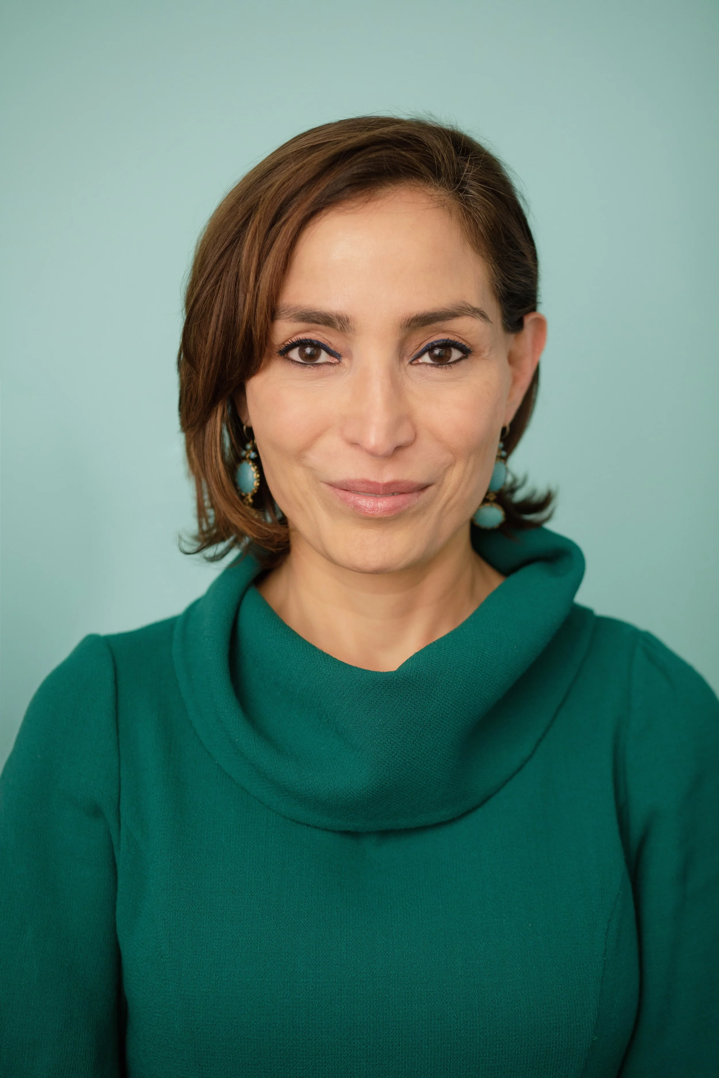 A woman with brown hair and brown eyes smiling, wearing a teal hoodie and turquoise earrings, against a light teal background.