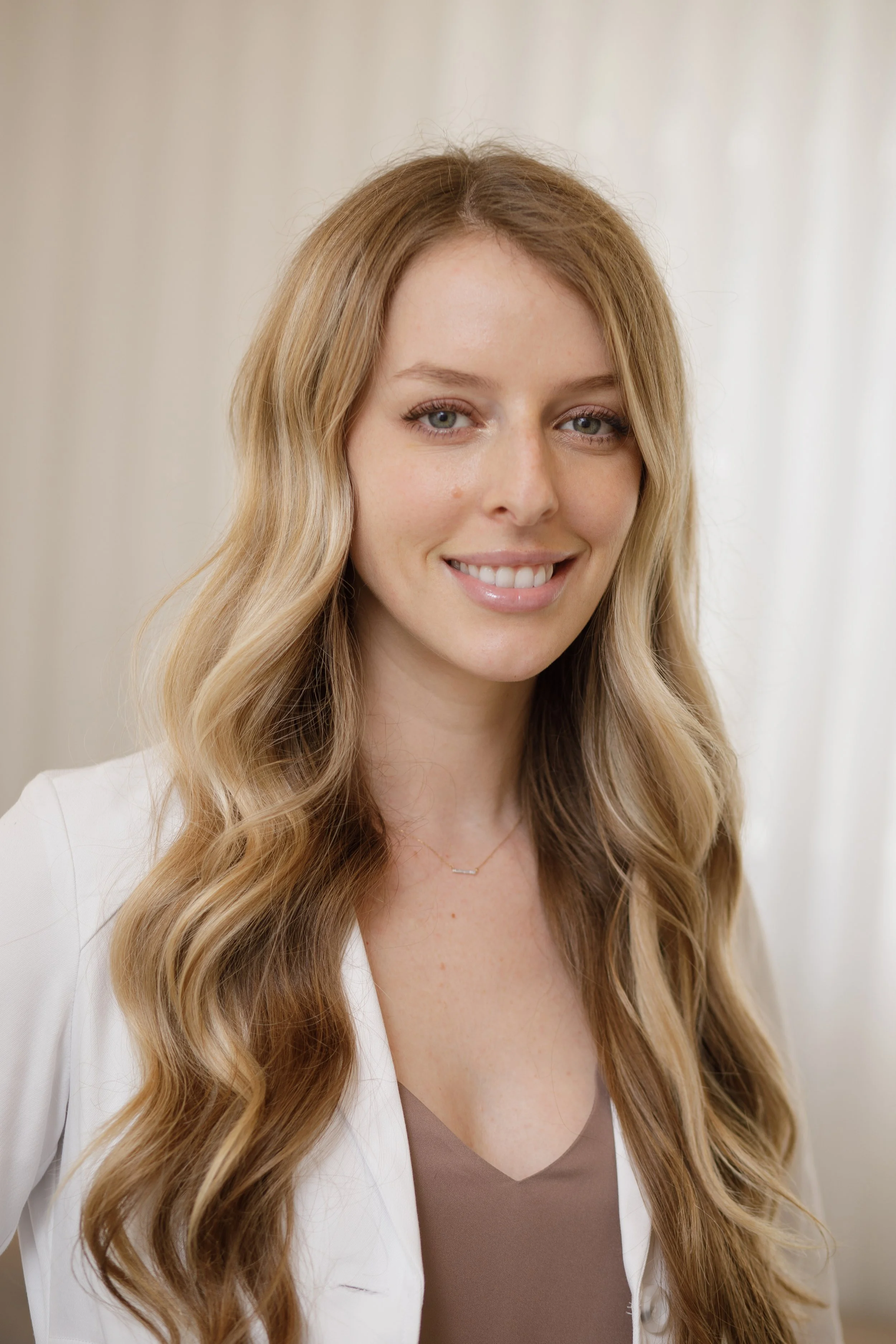 A smiling woman with long, wavy blonde hair wearing a white blazer over a beige top, standing against a light-colored background.