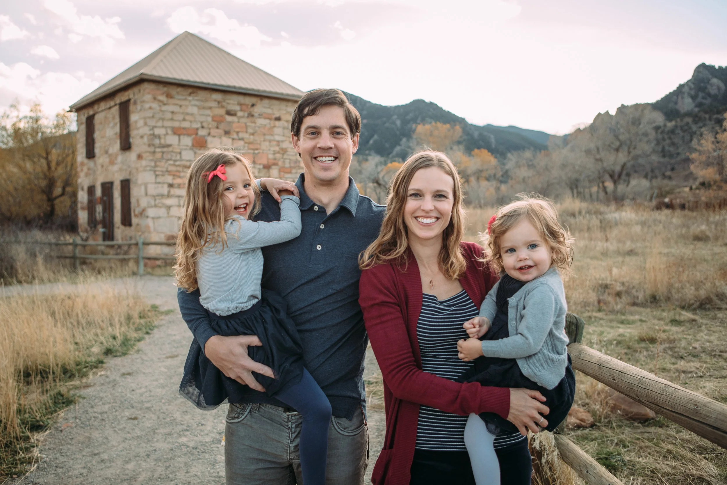 Family of four smiling outdoors with mountains and a stone building in the background, embracing each other.