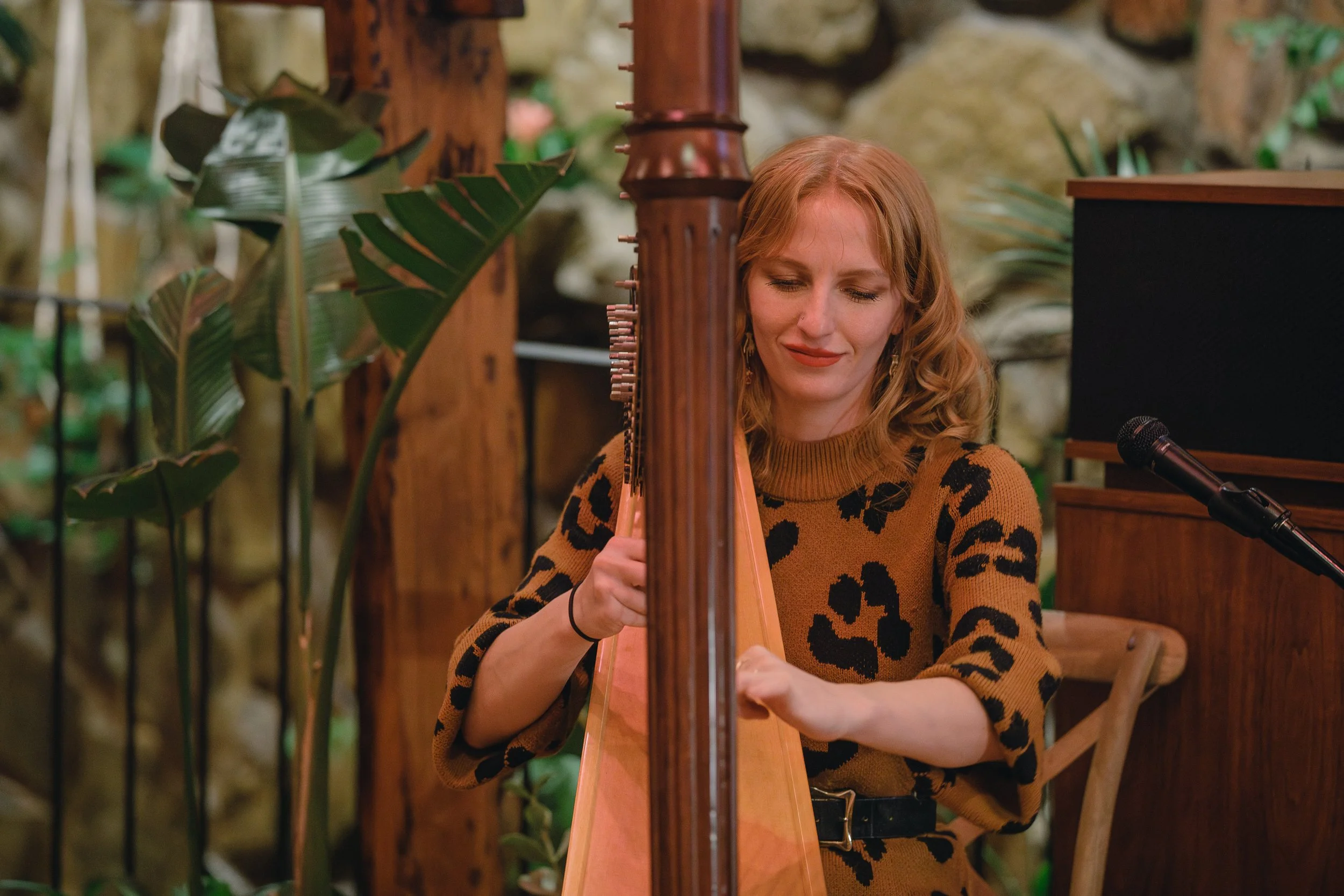 A woman with red hair wearing a leopard print sweater playing a harp in a cozy, plant-filled indoor setting.