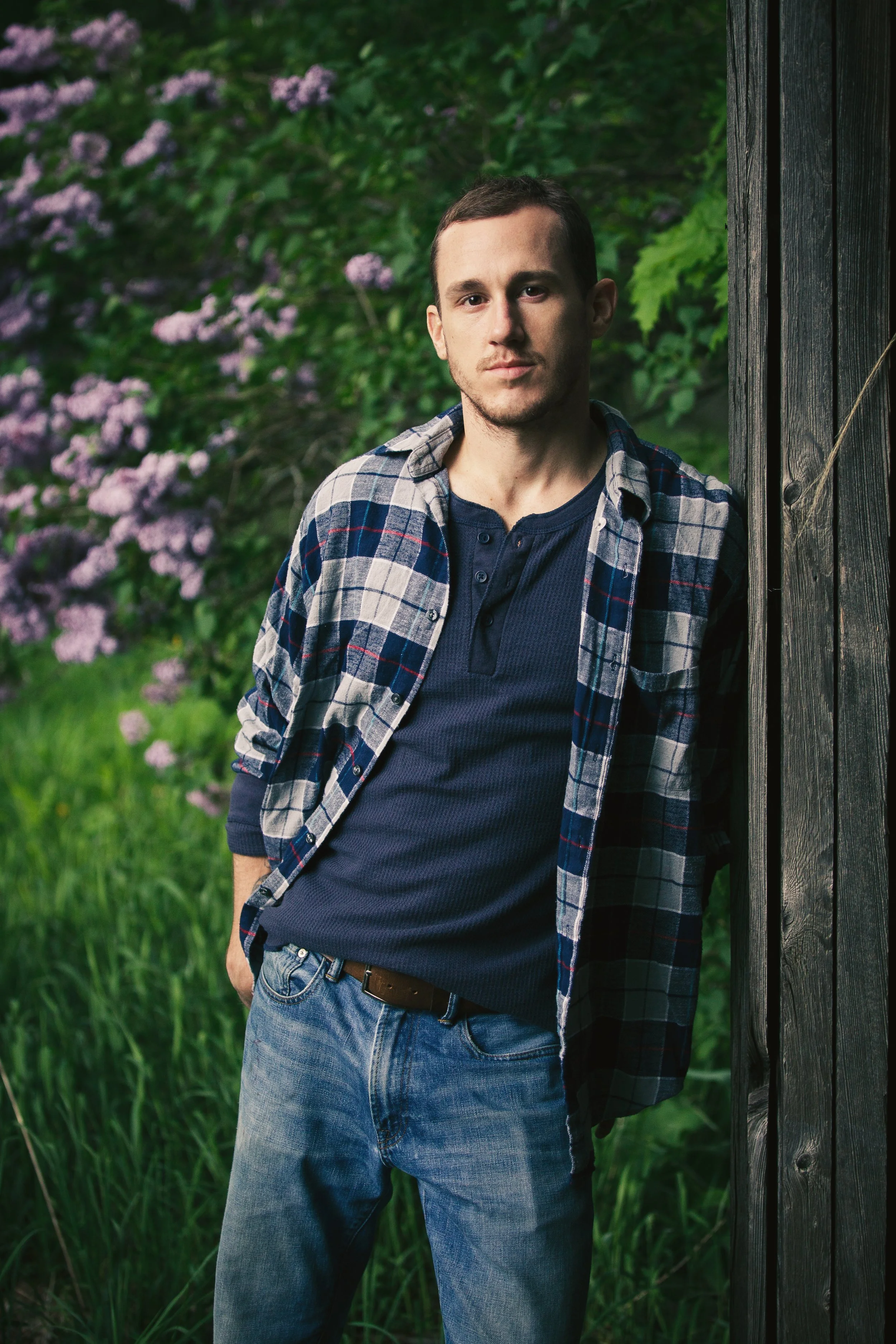 A young man with short brown hair and a slight beard standing outdoors next to a wooden post, wearing a blue T-shirt, a plaid shirt, and blue jeans, with a background of green foliage and purple flowers.