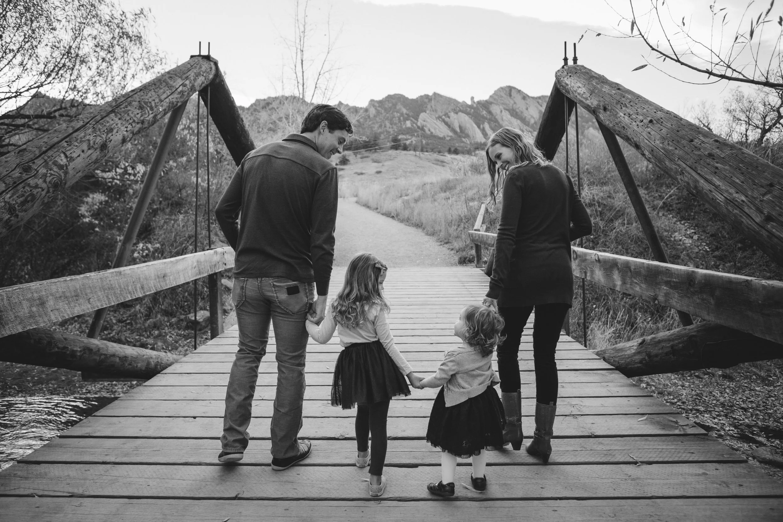 A family of four, including two young girls, stands on a wooden bridge holding hands in a natural outdoor setting with mountains in the background.