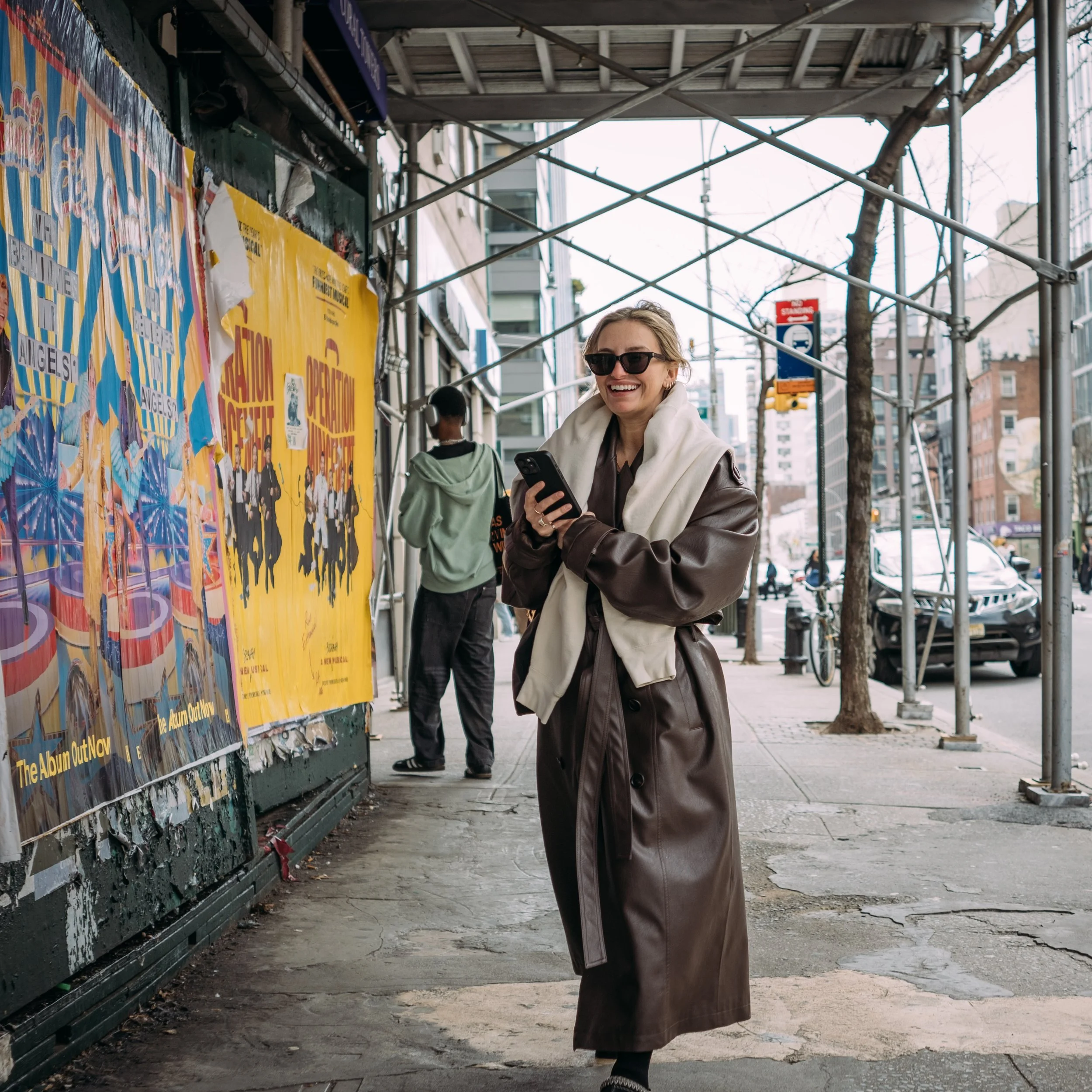 Smiling woman in sunglasses holding a phone while walking past a colorful poster on an urban street with scaffolding and a tree in the background.