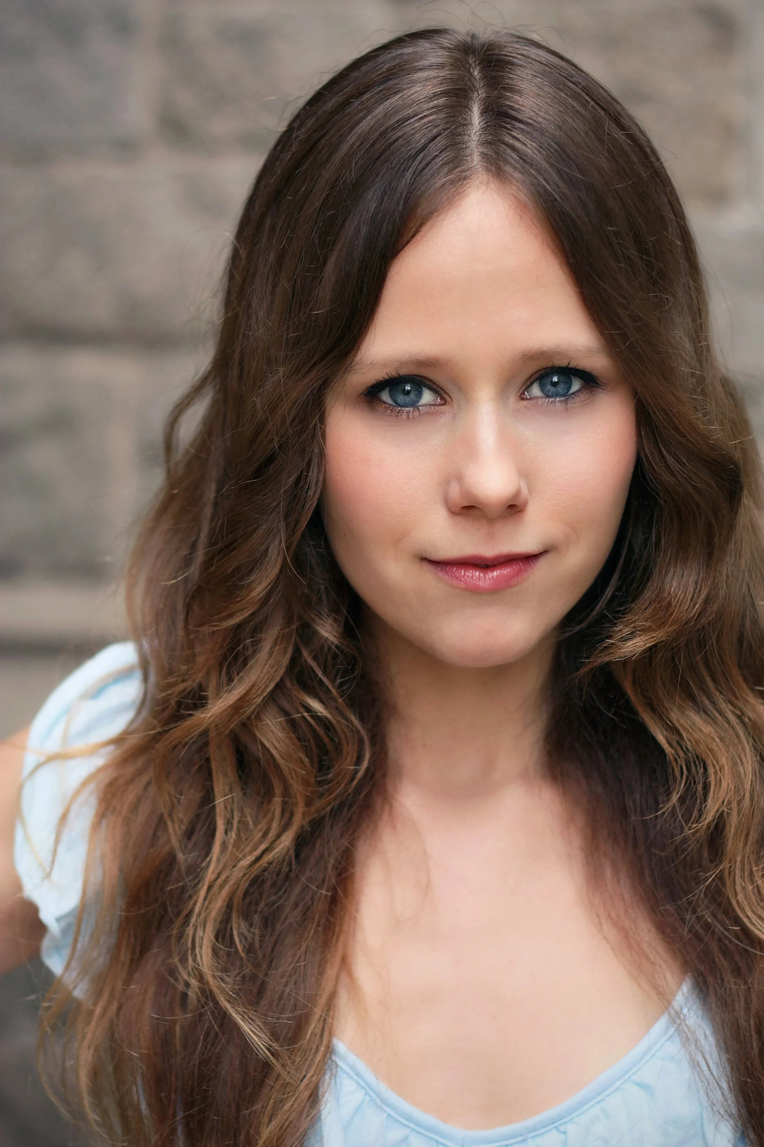 Close-up of a woman with long, wavy brown hair and blue eyes, wearing a light blue top, standing outdoors with a blurred brick wall background.