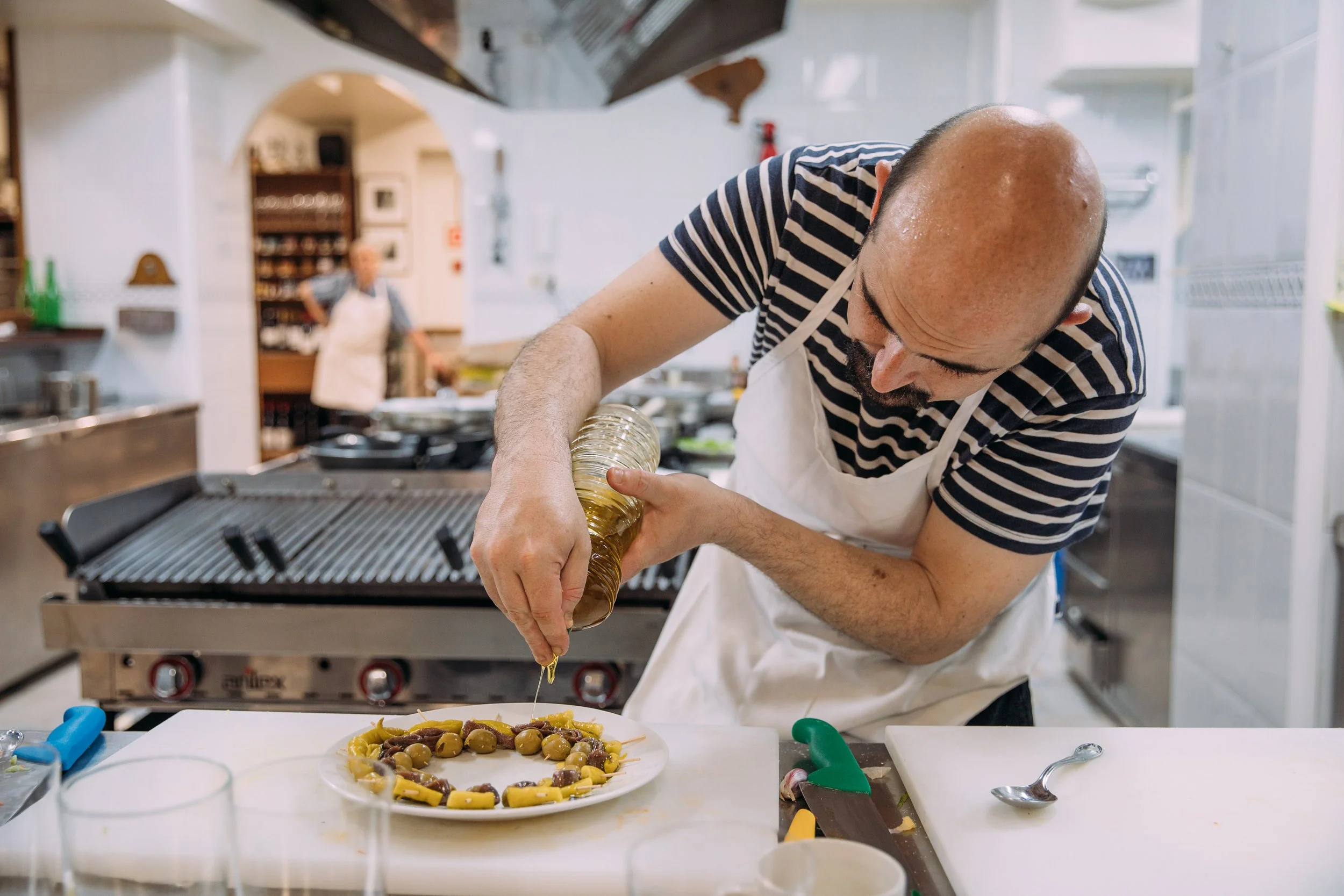 A man in a striped shirt and apron is pouring olive oil onto a plate of pasta and olives in a professional kitchen.