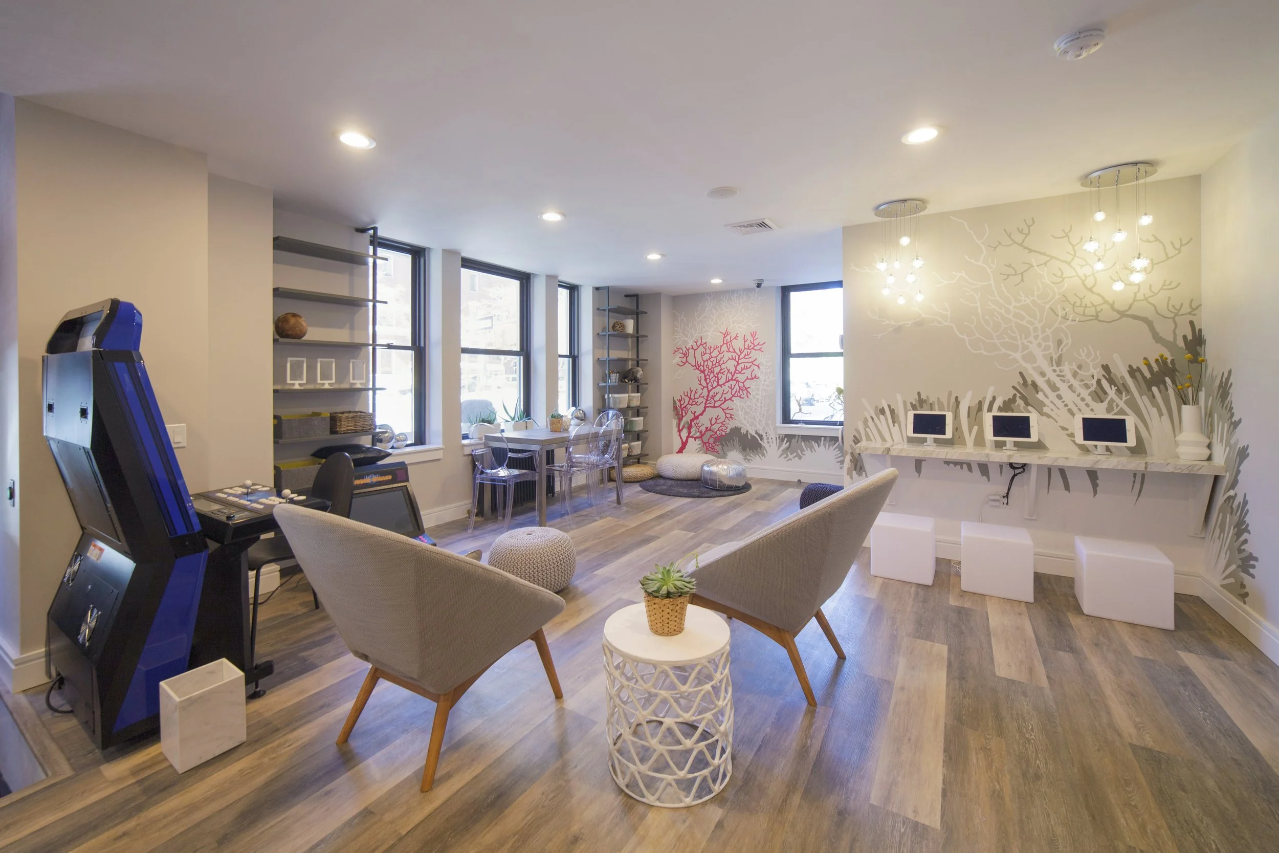 Interior of a modern lounge or waiting area with large windows, wall art featuring coral and tree designs, white seating, tables with decorative items, and a row of small computers or displays on a white desk.