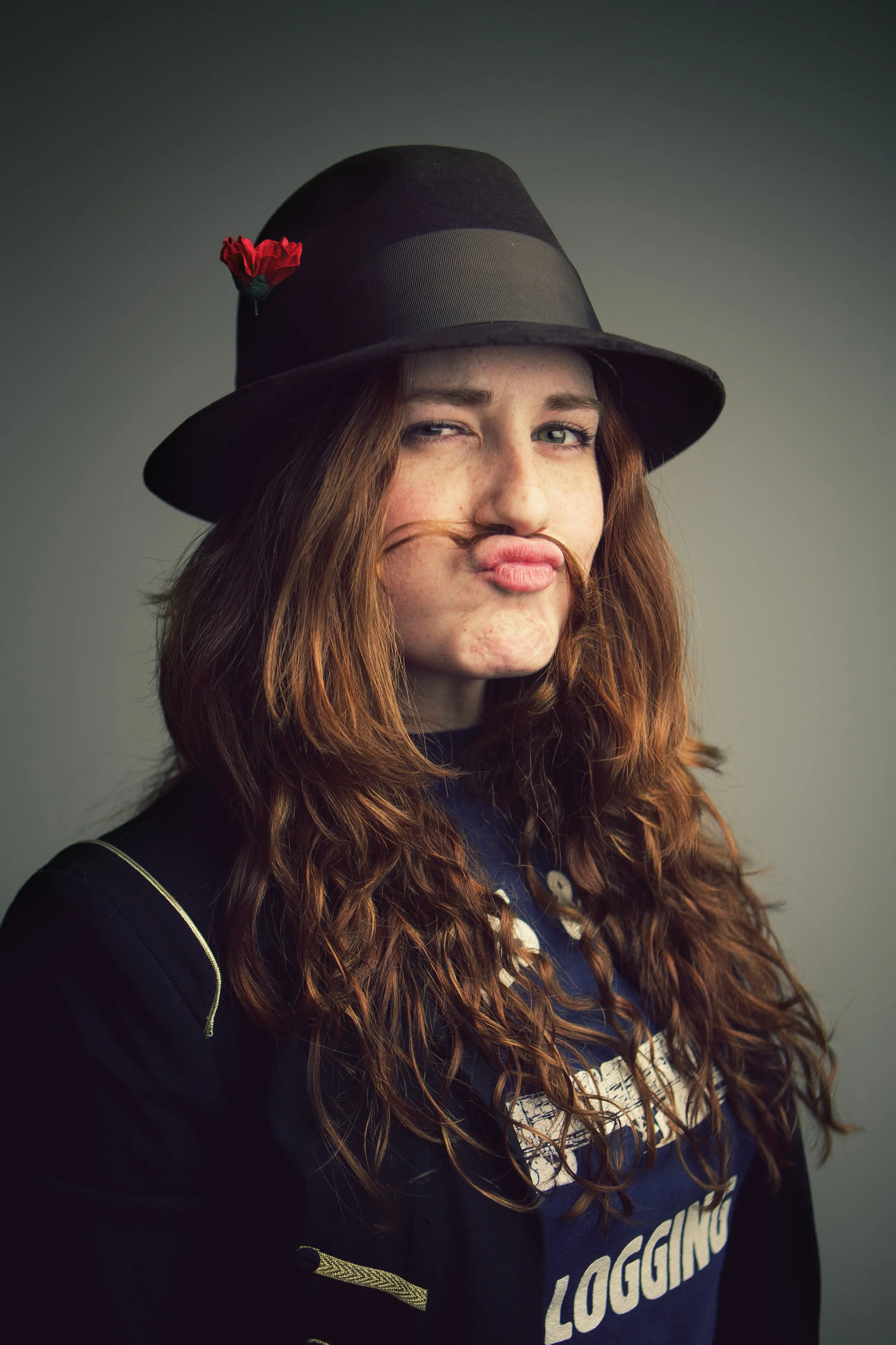 A young woman with red hair, wearing a black fedora hat with a red flower, making a pouting face, wearing a dark jacket with gold accents and a blue shirt underneath with white text.