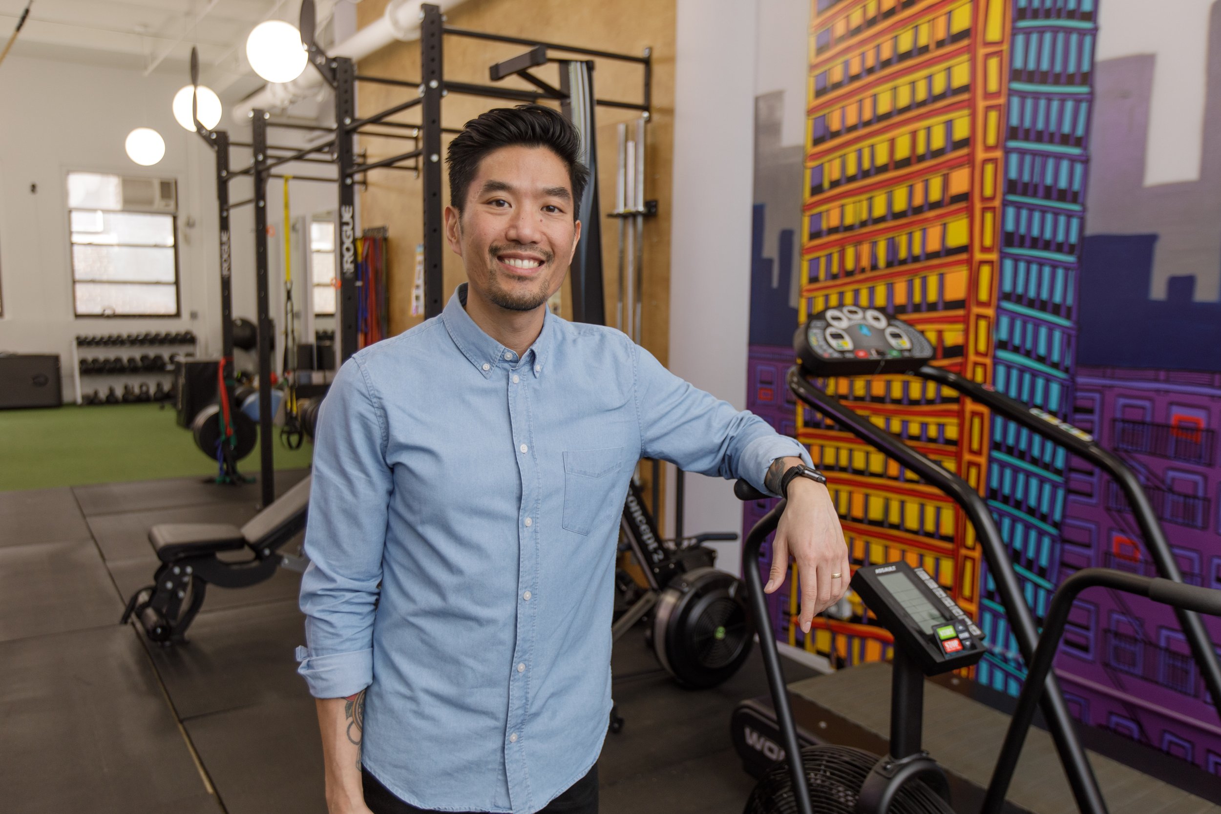 A smiling man in a light blue shirt standing in a gym next to exercise equipment and a colorful mural.