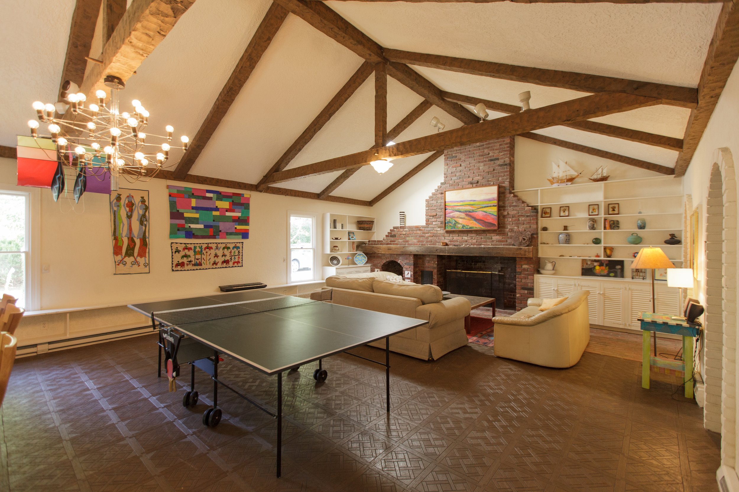 A spacious living room with a high vaulted ceiling featuring exposed wooden beams and white ceilings. The room has a brick fireplace with a colorful landscape painting above it. There are large windows allowing natural light, with art and decorative 