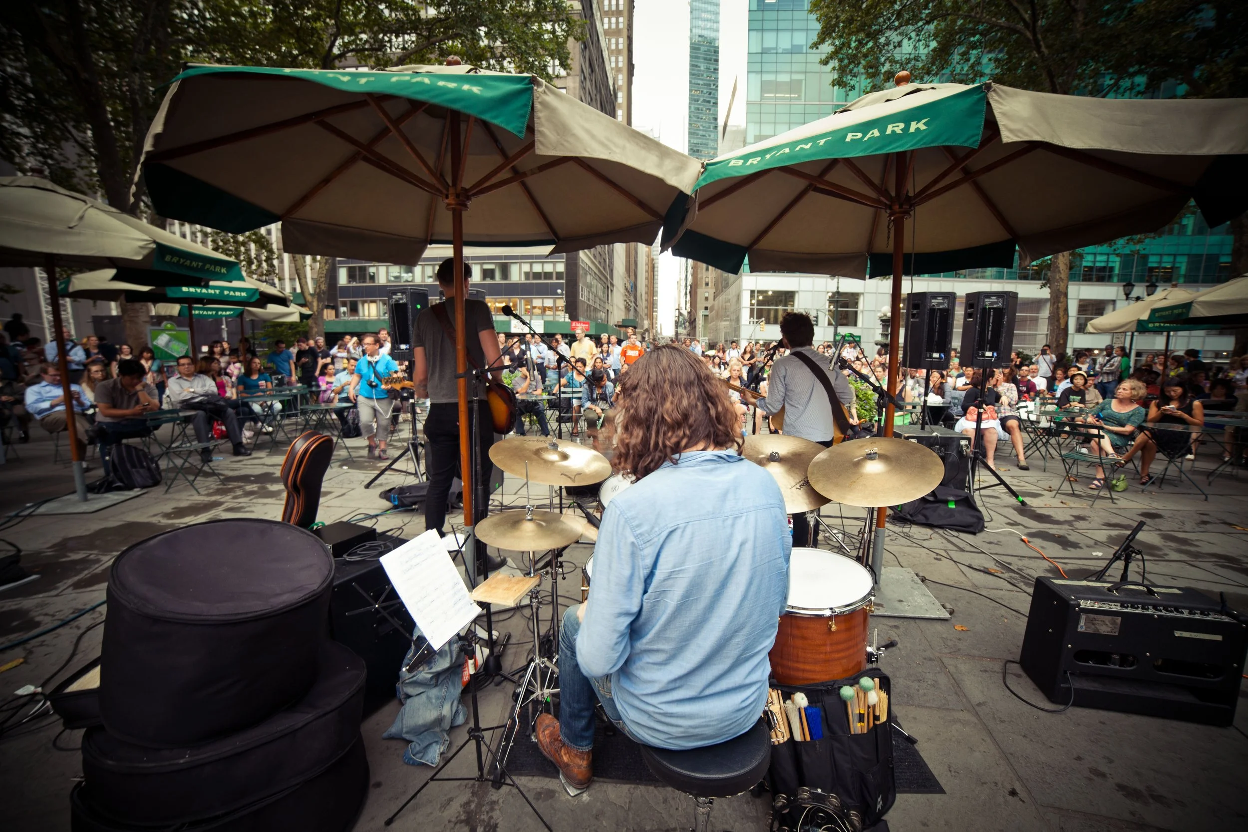 A band performing on an outdoor stage in Bryant Park during daytime, with an audience sitting under umbrellas and tall buildings in the background.