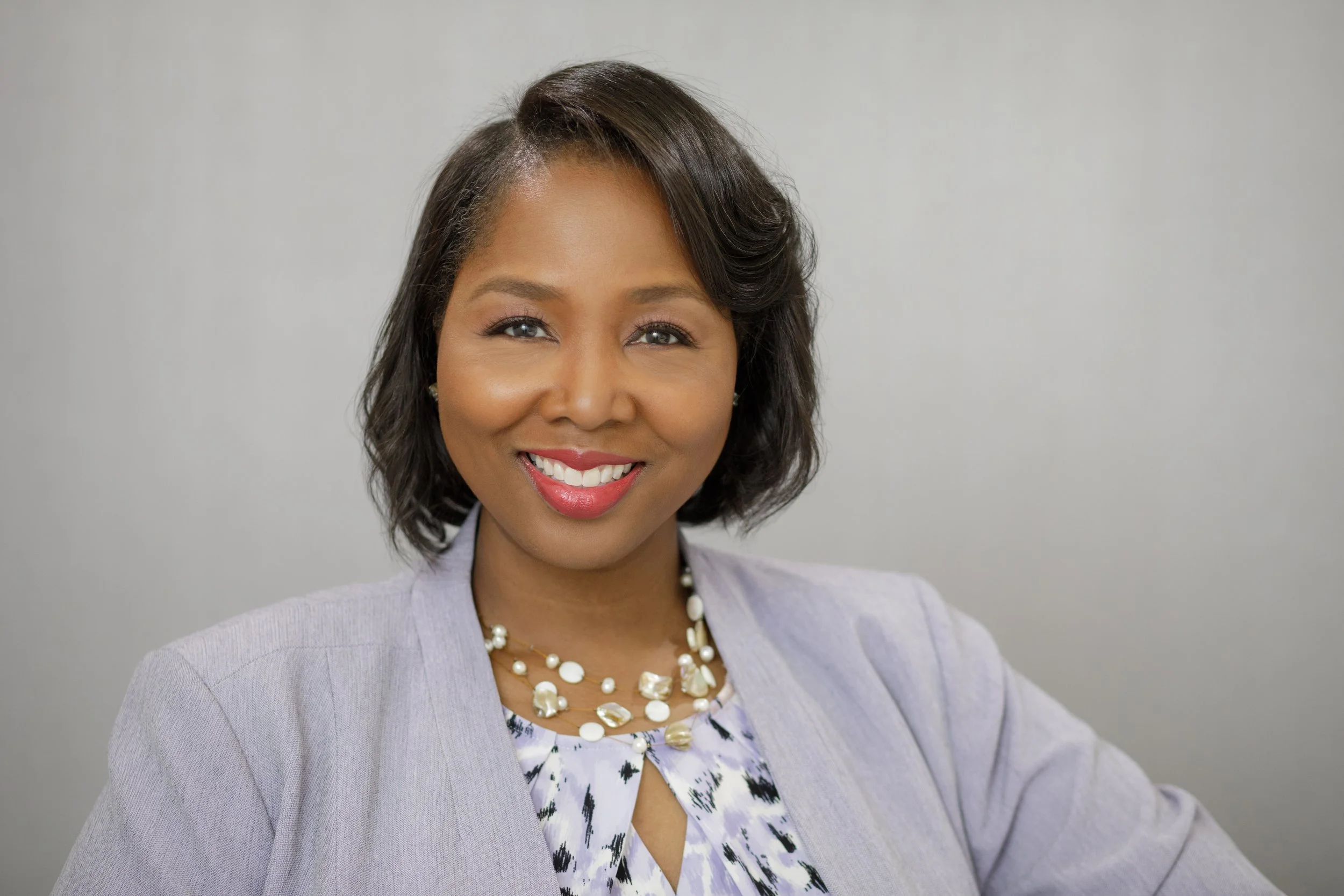 A smiling woman with shoulder-length dark hair styled with a side part in a professional setting, wearing a light gray blazer over a printed blouse, accessorized with a pearl necklace and earrings, standing against a plain gray background.