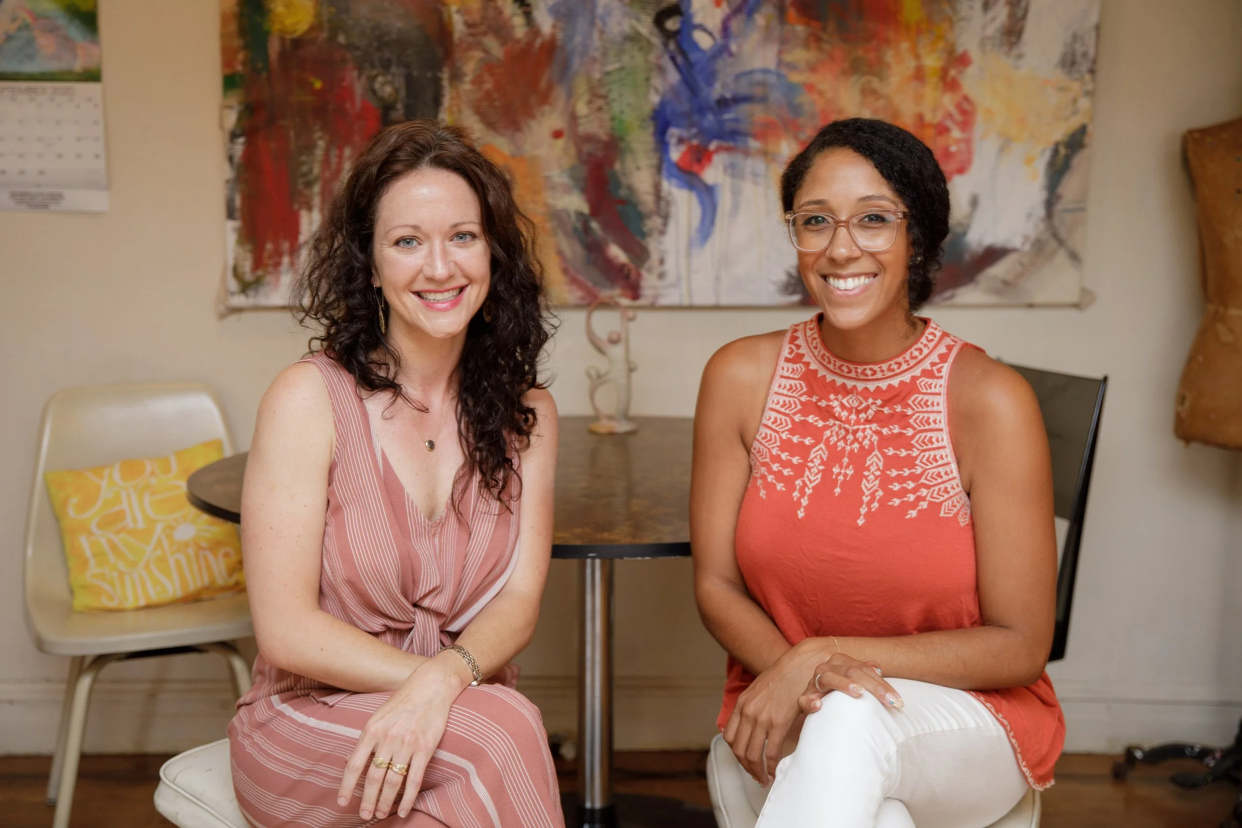 Two women sitting at a small round table in a cozy indoor space. The woman on the left has light skin, brown curly hair, and is wearing a pink striped dress with a gold necklace. The woman on the right has darker skin, short dark hair, and glasses, w