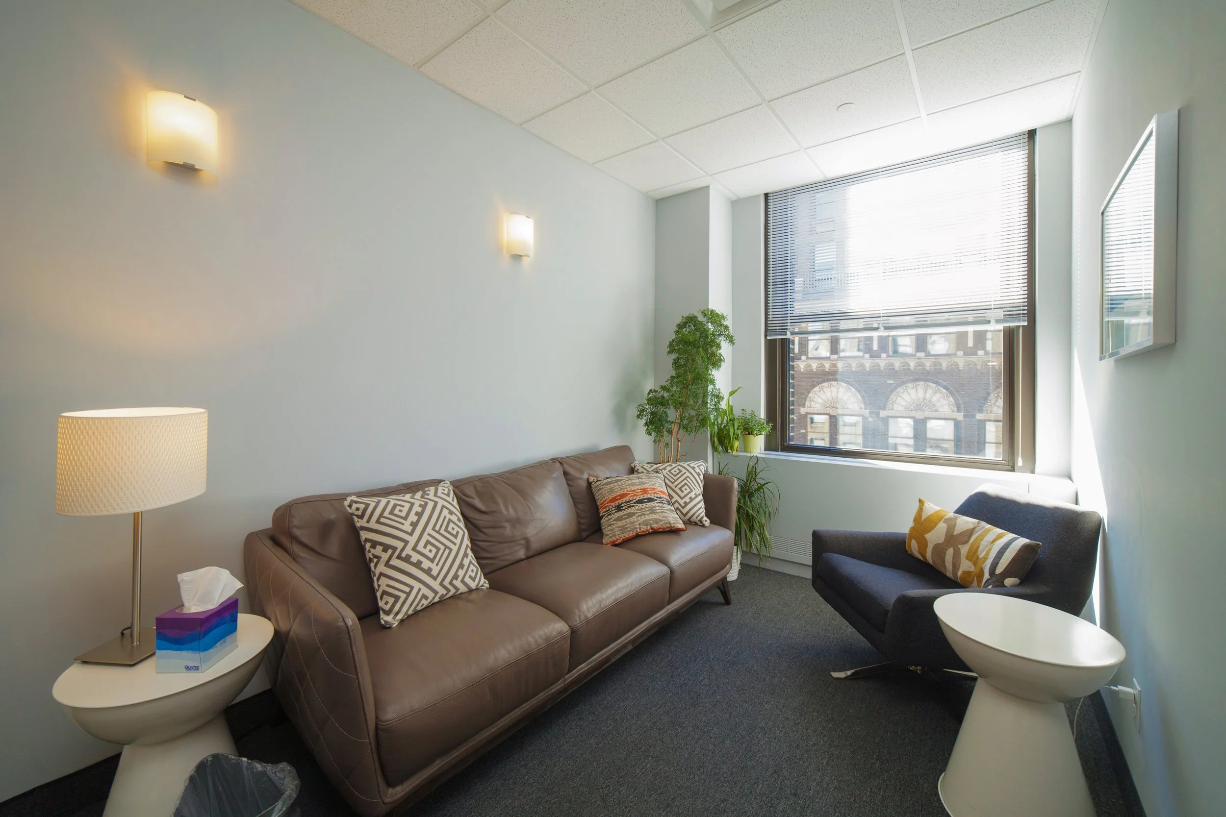 A cozy office or lounge area with a light blue wall, sofa with patterned pillows, a black chair with a decorative pillow, a white side table with tissues, a small white round table, a tall floor lamp, and a window with blinds revealing a historic bui
