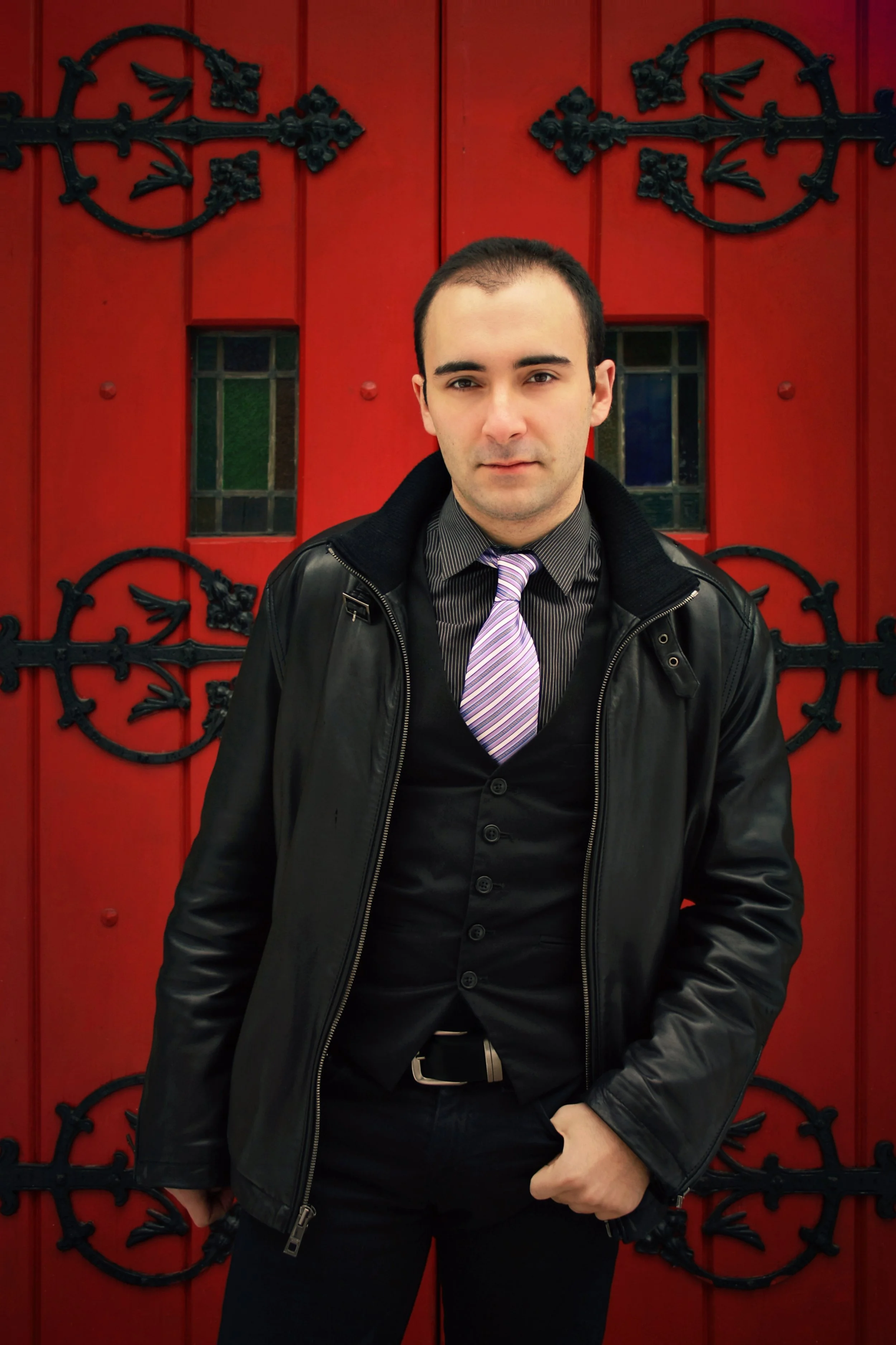 A young man with dark hair, wearing a black leather jacket, black shirt, purple striped tie, and black pants, standing in front of a red door with ornate black ironwork.