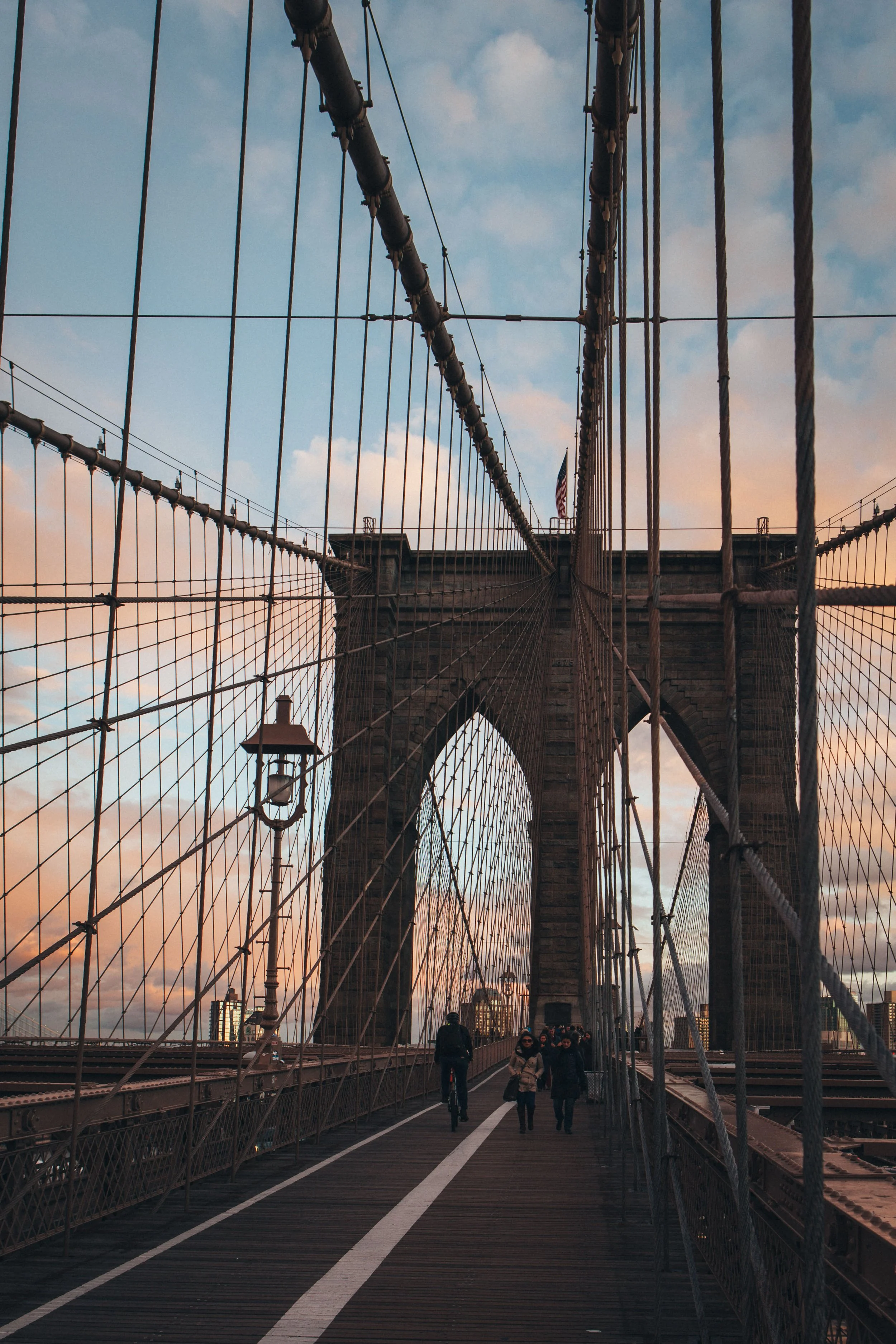 View of the Brooklyn Bridge in New York City during sunset with people walking across the pedestrian walkway, featuring cables and stone towers with an American flag.