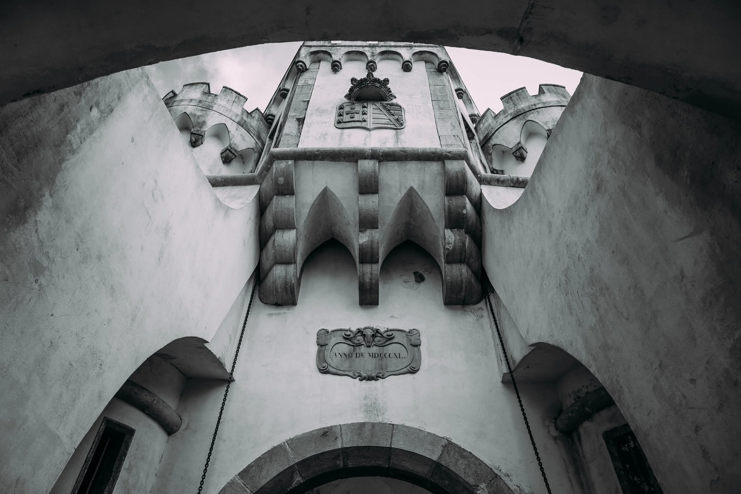 A black-and-white photograph of a castle-like structure's interior with a monument or shield featuring a coat of arms and an inscription reading "Anno de MDCXXCL". The view is from below, looking upward through a curved archway.