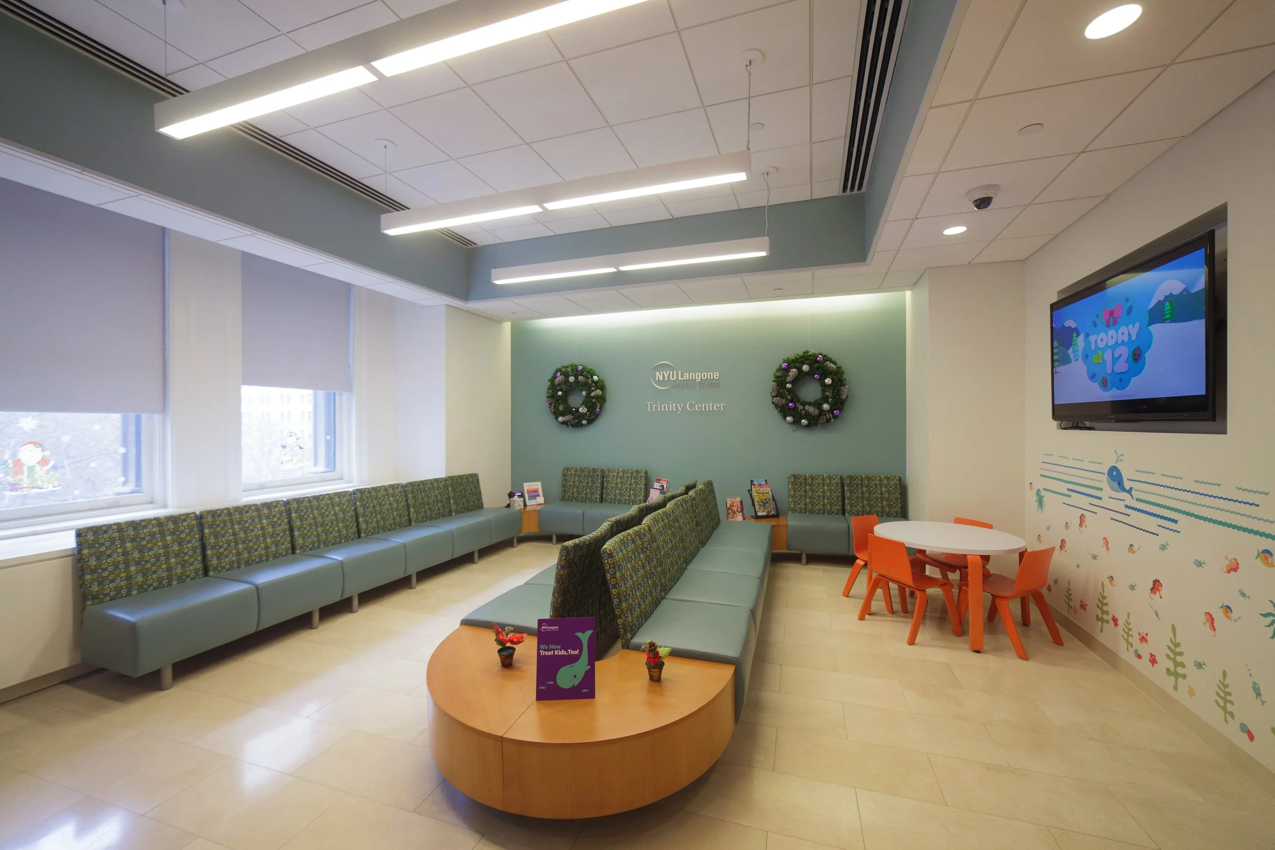 Lounge area with green cushioned seating, small round tables, Christmas wreaths on the wall, a television displaying a children's show, and various decorative elements in a hospital or clinic waiting room.