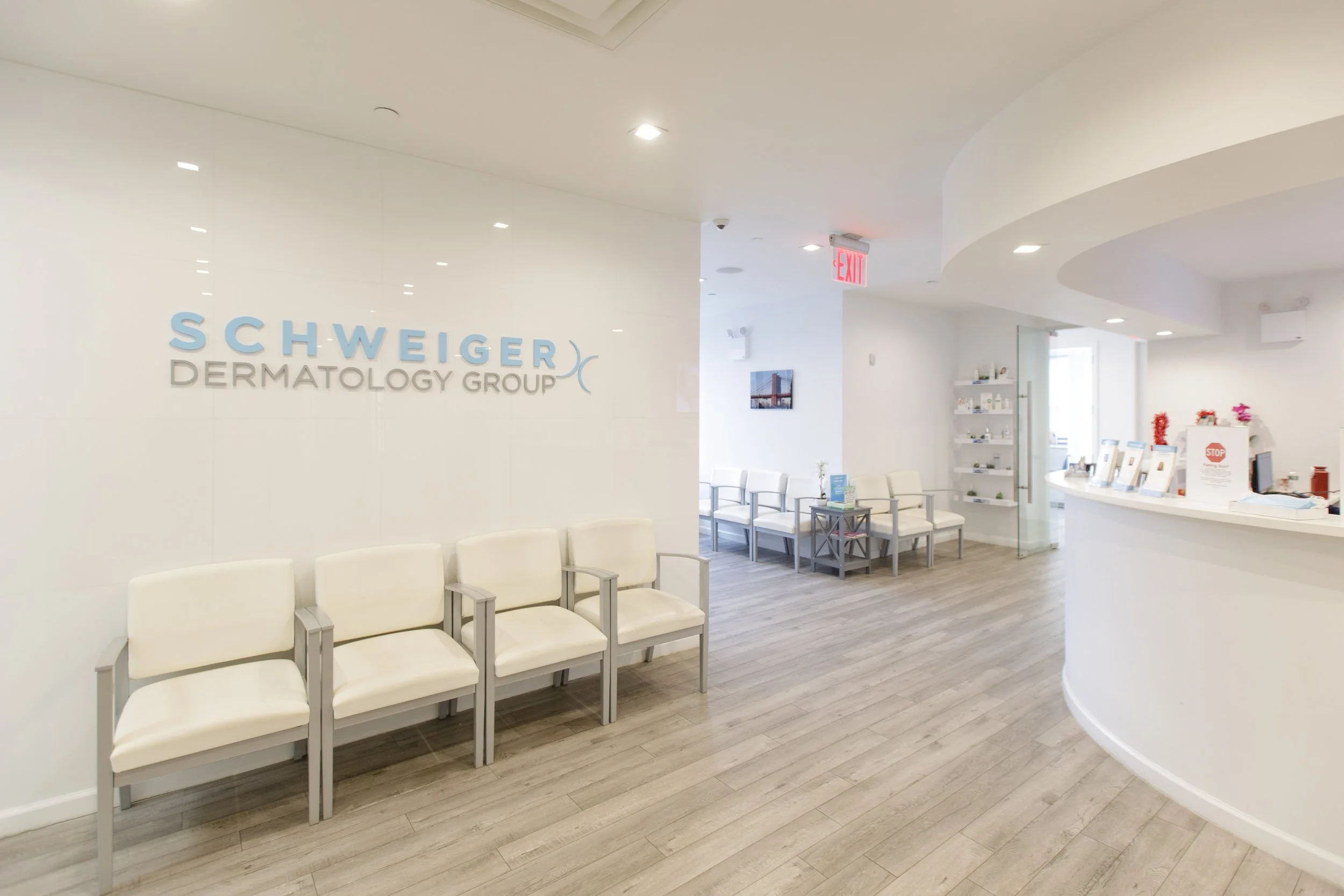 The interior of a dermatology office with a waiting area, white walls, beige chairs, a reception desk, and signage that reads 'Schweiger Dermatology Group' on the wall.