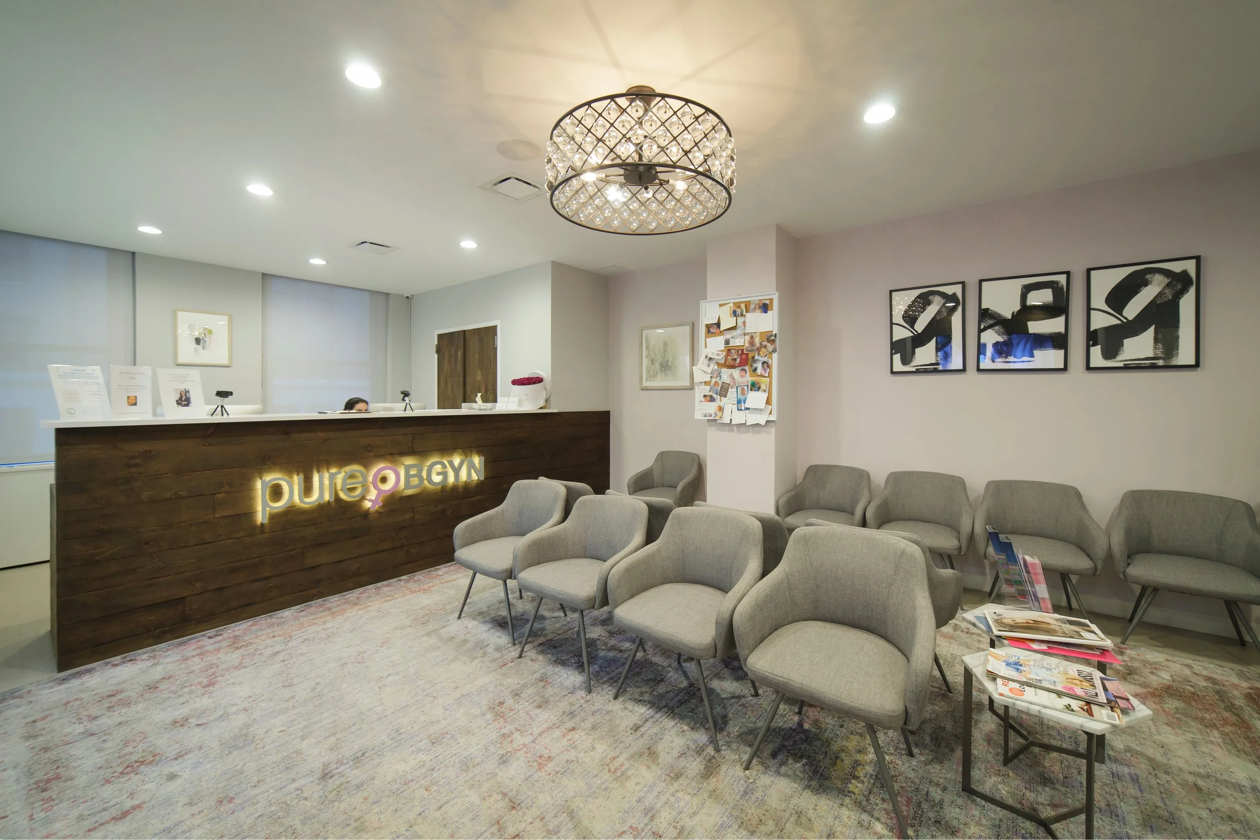 Reception area of a clinic or office with a wooden front desk, a lit sign reading 'pure OBGYN', chairs for waiting, and modern artwork on the walls.