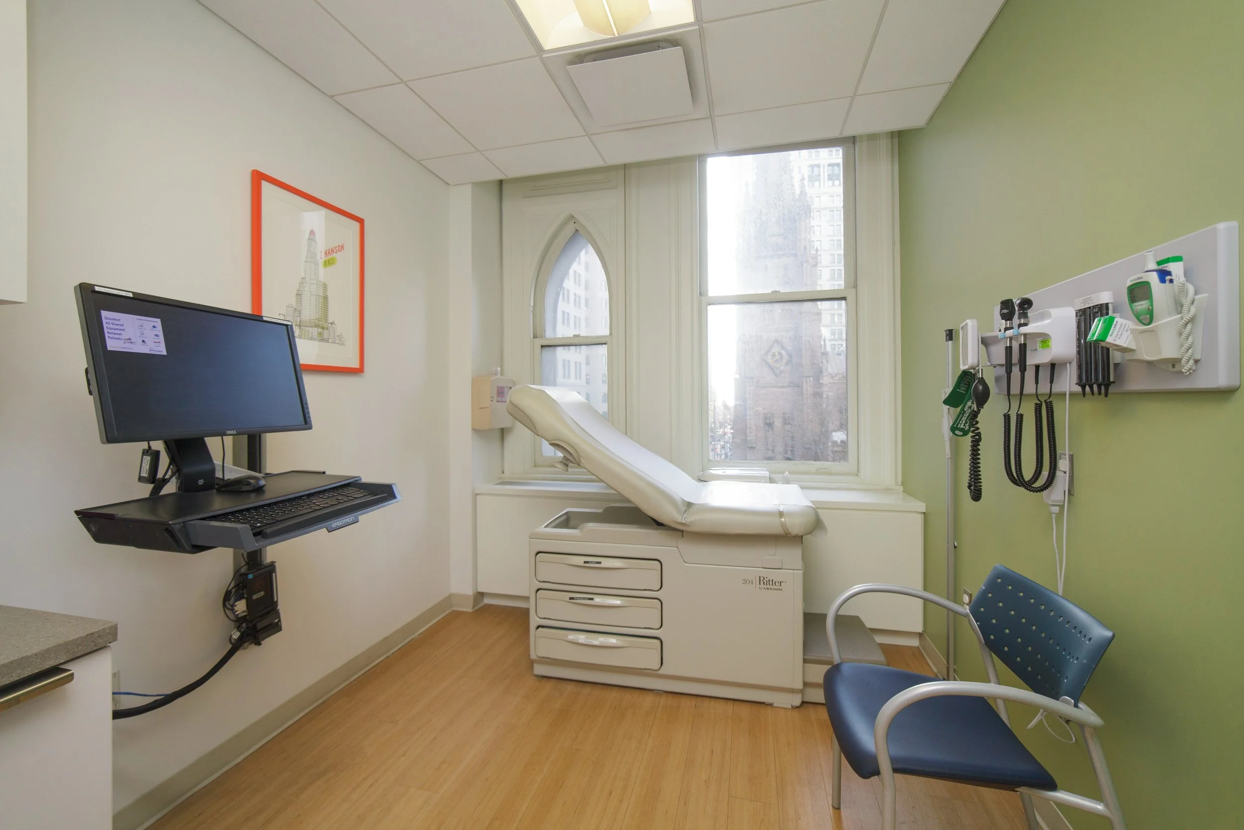 A medical examination room with a patient bed near a window, a blue chair, a wall-mounted medical tools panel, a computer monitor on a movable arm, artwork on the wall, and cabinets underneath the bed.