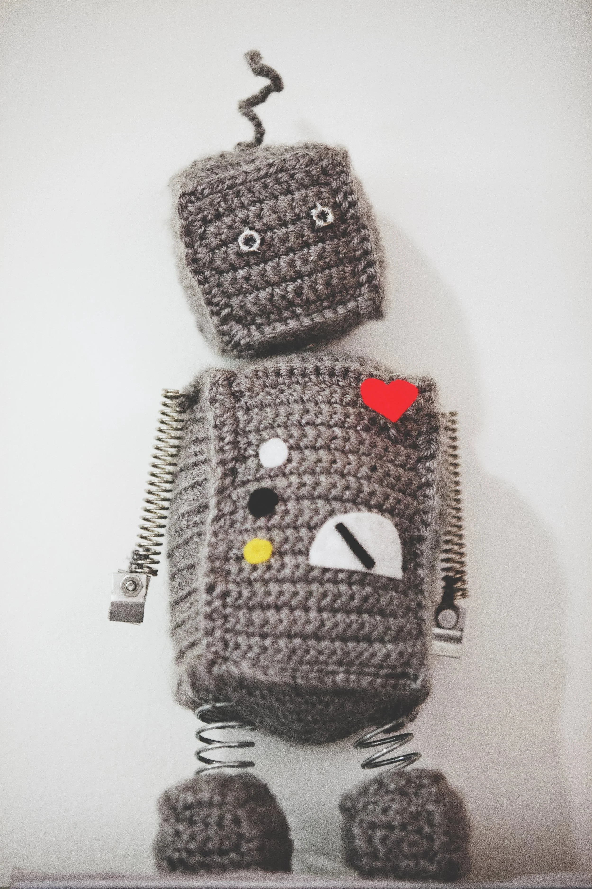 Close-up of a plush toy robot made with gray knitted fabric, featuring small embroidered eyes, a red heart on its chest, and a zipper on its arm, set against a plain white background.