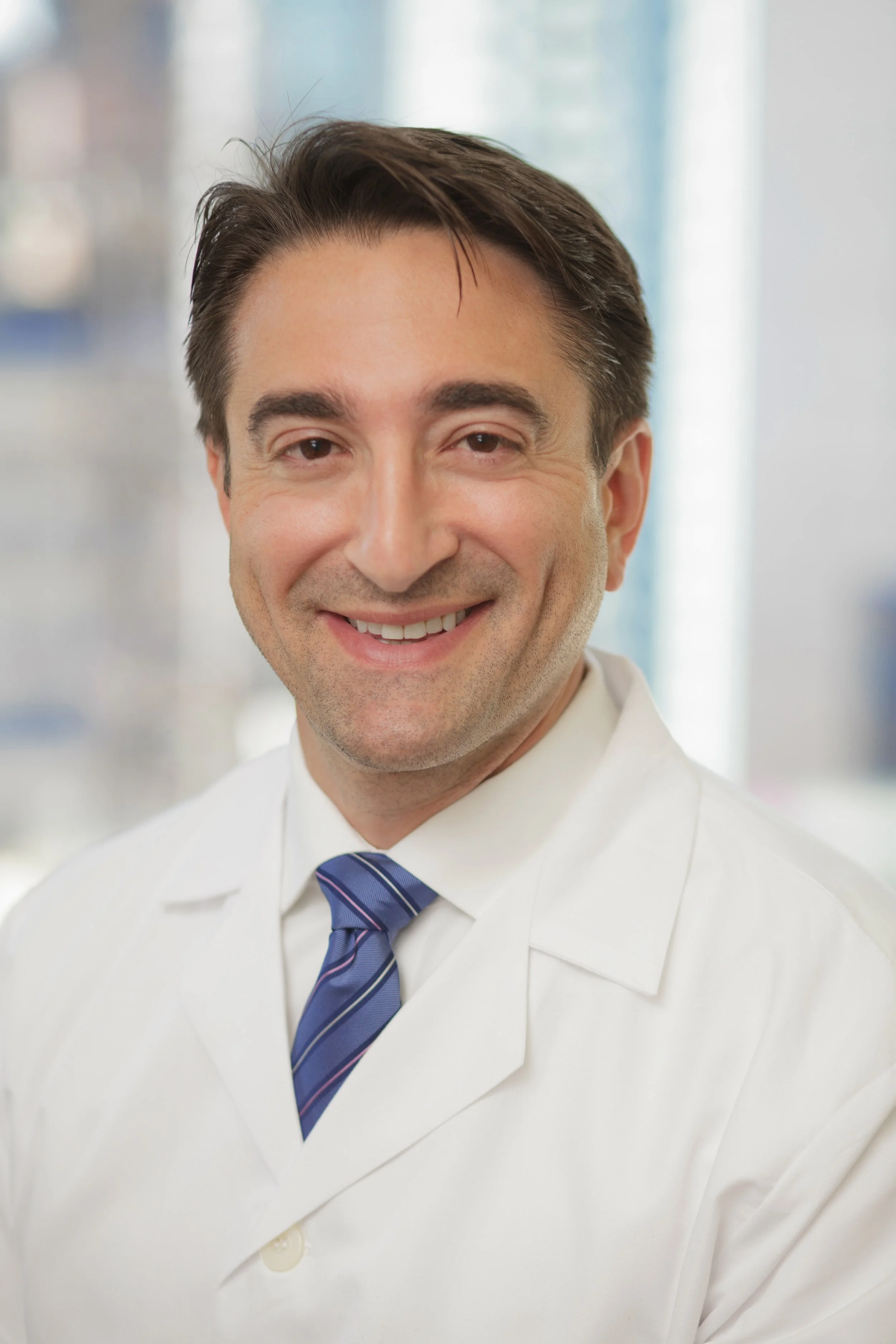 Smiling man in a white lab coat, blue striped tie, in a professional setting with natural light and blurred background.
