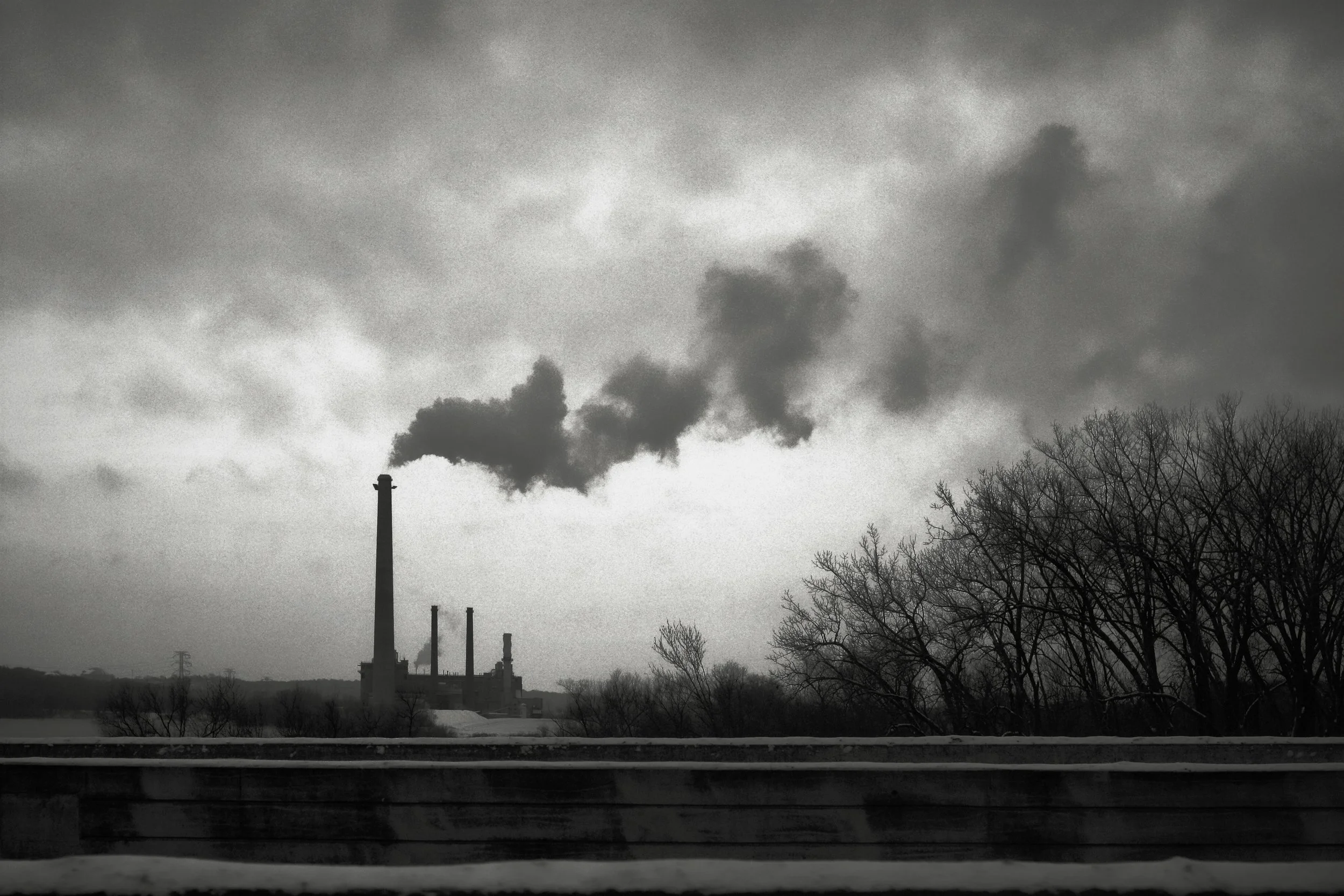 Black and white photo of an industrial factory with tall smokestacks emitting dark smoke into the sky, with leafless trees in the foreground and clouds.