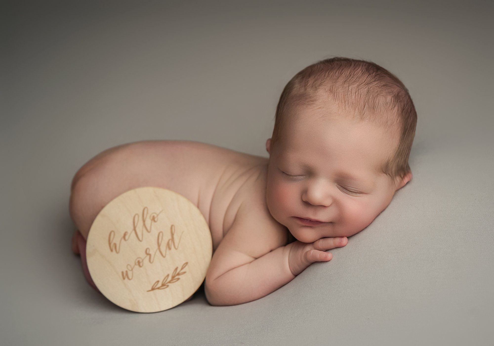 A newborn baby sleeping on a soft surface, with a round wooden sign reading 'hello world' placed on their back.