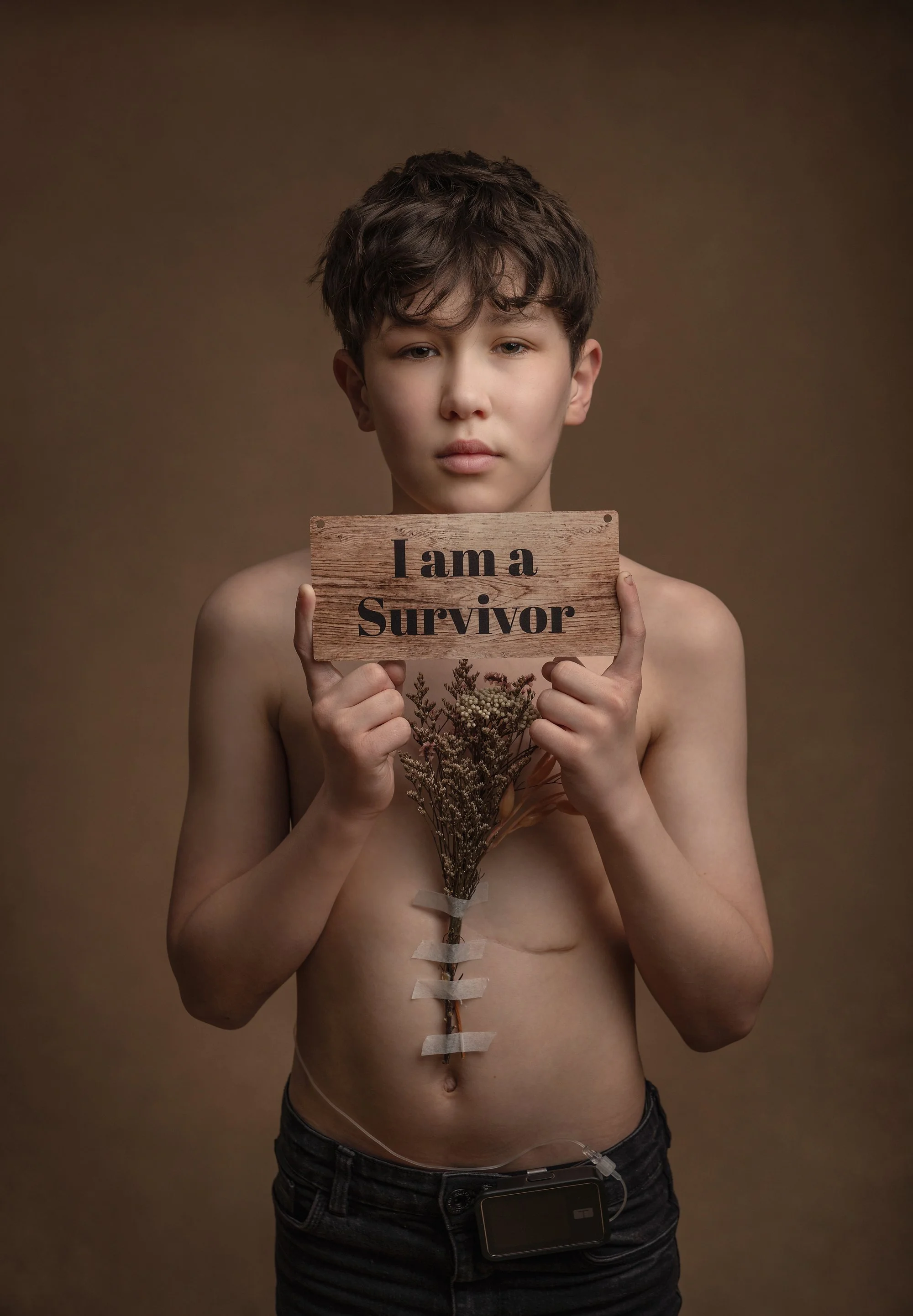 A shirtless young boy with a serious expression holding a wooden sign that reads 'I am a Survivor.' A natural bouquet of dried flowers is taped to his chest photographed at kath evans photography studio talgarth brecon mid wales