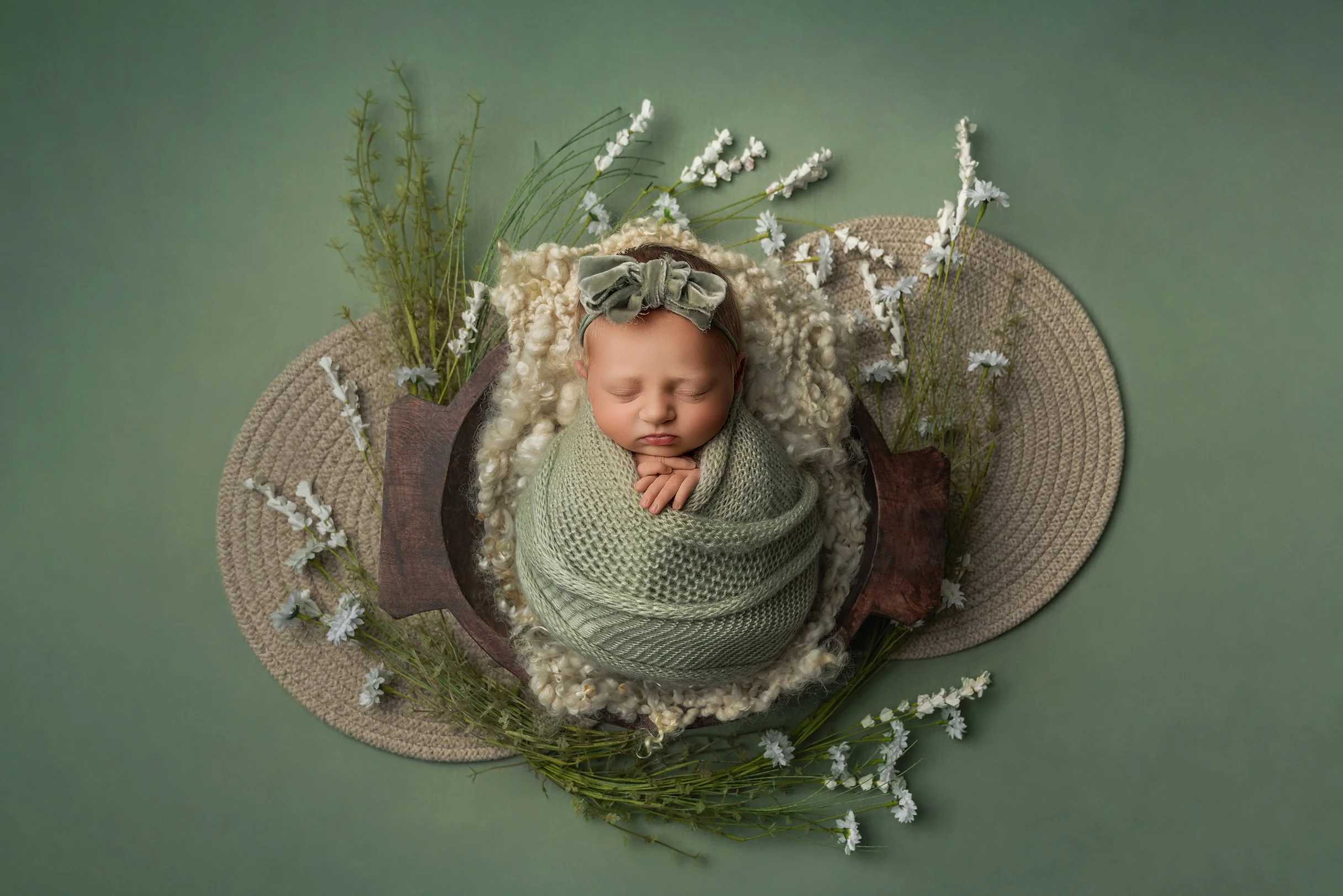 A newborn baby with curly blond hair, peacefully sleeping, wrapped in a green knit blanket and wearing a matching green bow headband, surrounded by white flowers and greenery on a soft green background.