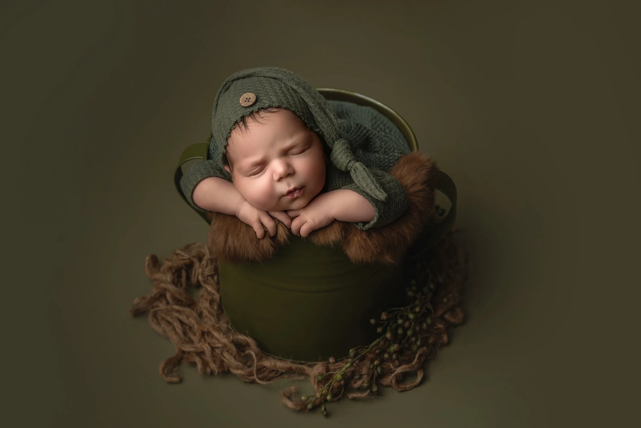A sleeping baby in a green hat and matching outfit, resting with hands under chin, inside a green bucket lined with brown fur, surrounded by a cluster of twine and small berries on a green background.