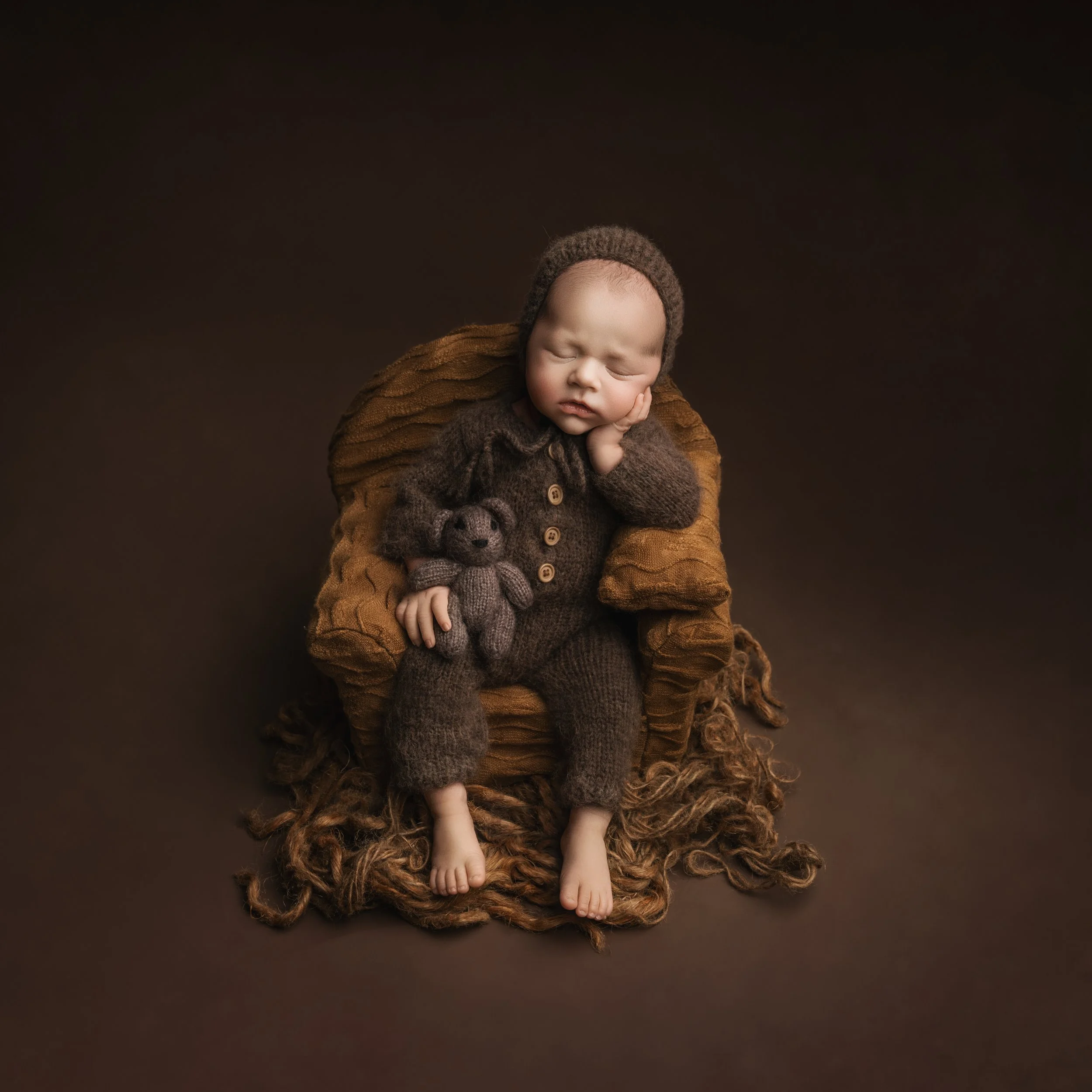 Young baby dressed in brown knitted outfit with a teddy bear, sitting on a patterned cushion, against a dark brown background.