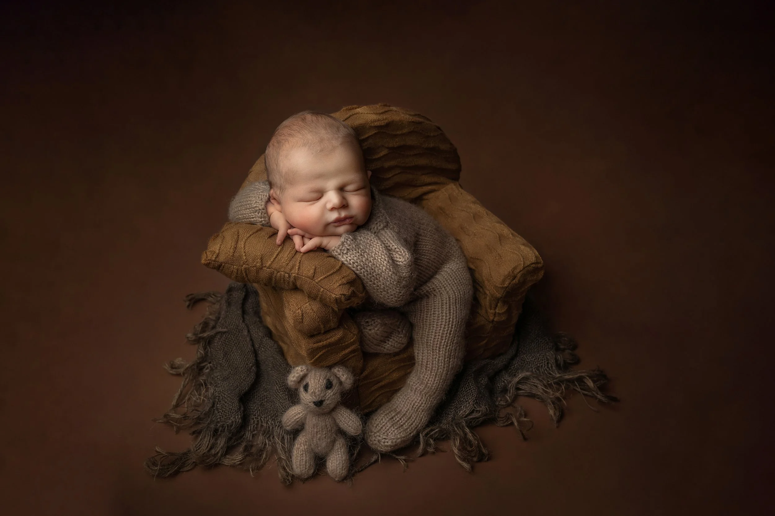 Sleeping baby seated on a brown pillow with a small knitted teddy bear in front, all on a dark brown background.