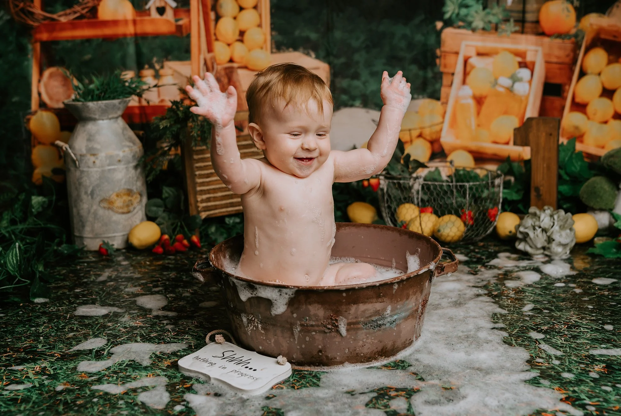 A smiling toddler sitting in a rustic brown tub, covered in soapy suds, with arms raised joyfully during a bath time photo photographed at kath evans photography studio talgarth brecon mid wales