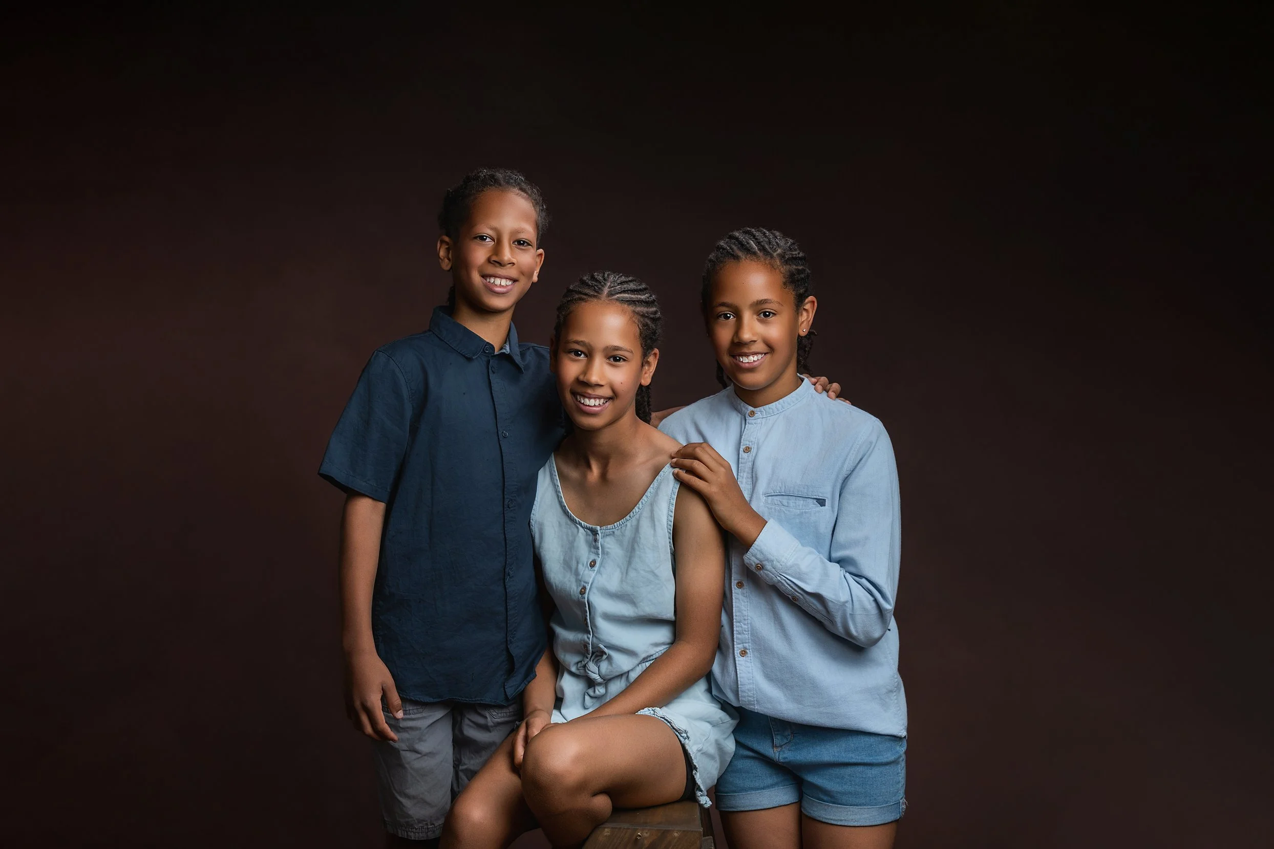 Three young girls with dark hair and dark skin, smiling and posing together against a dark brown background, one seated and two standing behind her with their arms around each other.