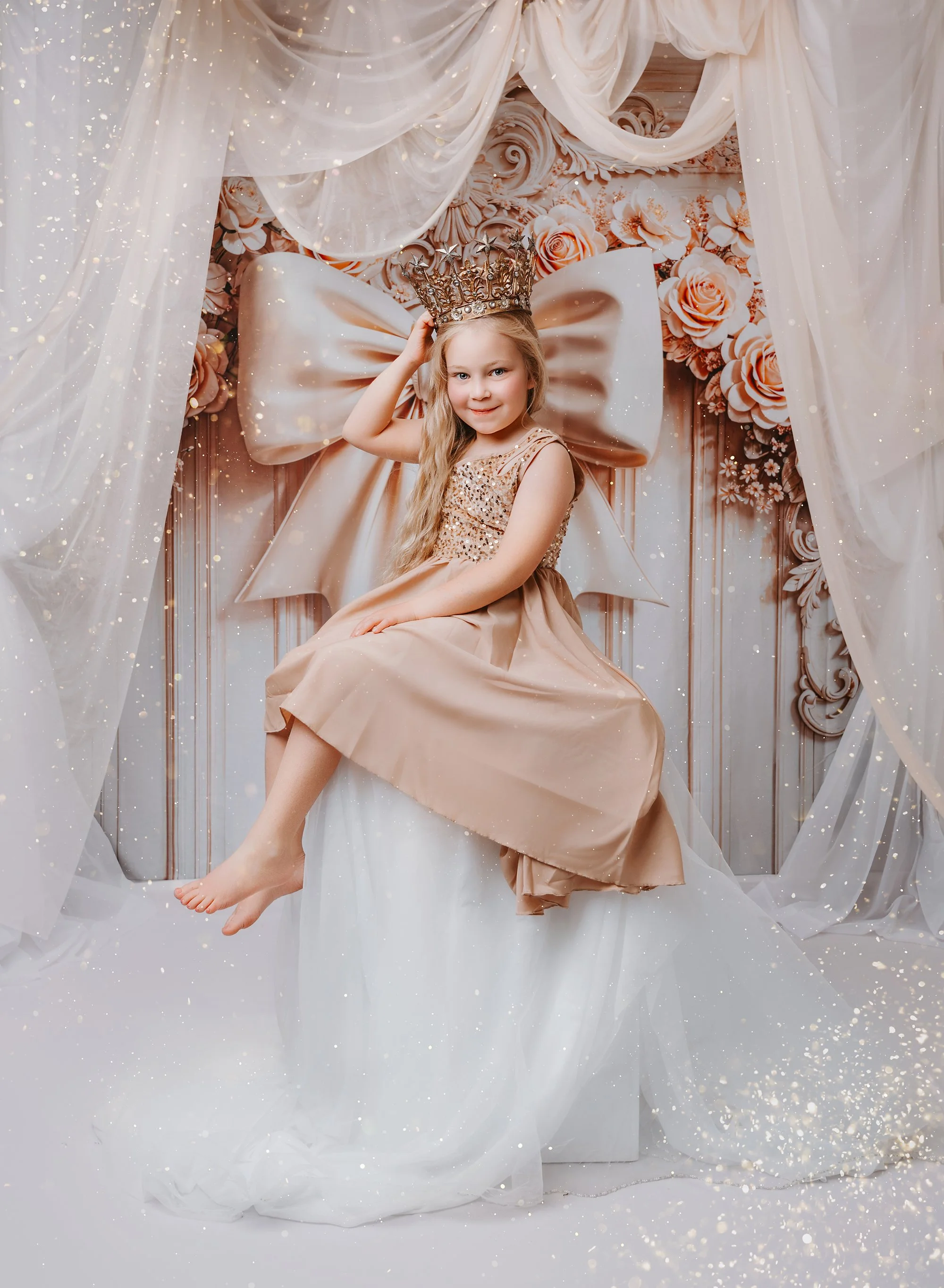 A young girl with long blonde hair, wearing a beige dress with sequins, sitting on a white stool surrounded by white drapes, behind a large golden bow, a crown, and floral decor, with gold glitter effects.
